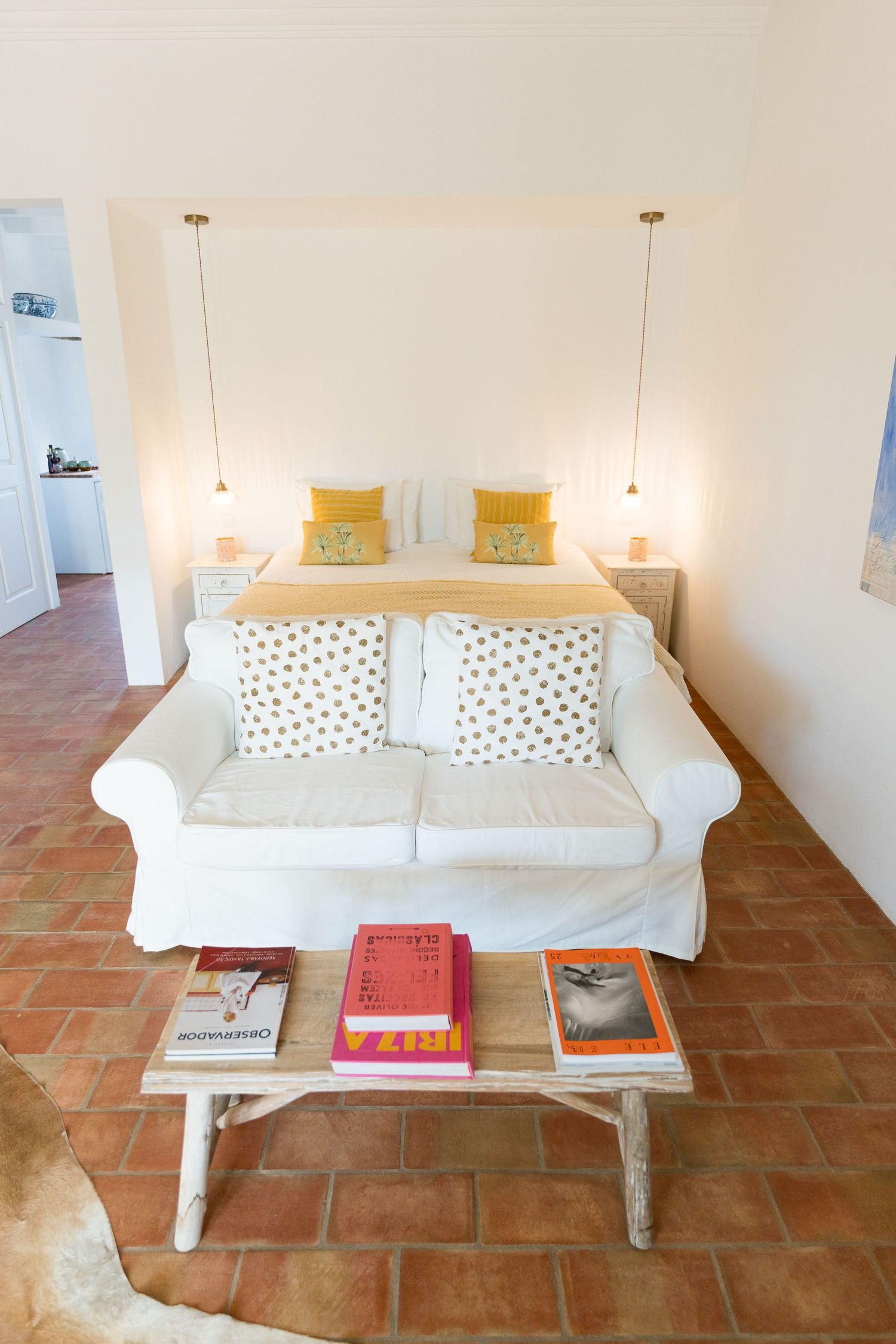 White sofa with polka dot pillows and yellow cushions beside a wooden coffee table with books in a terracotta-floored room.