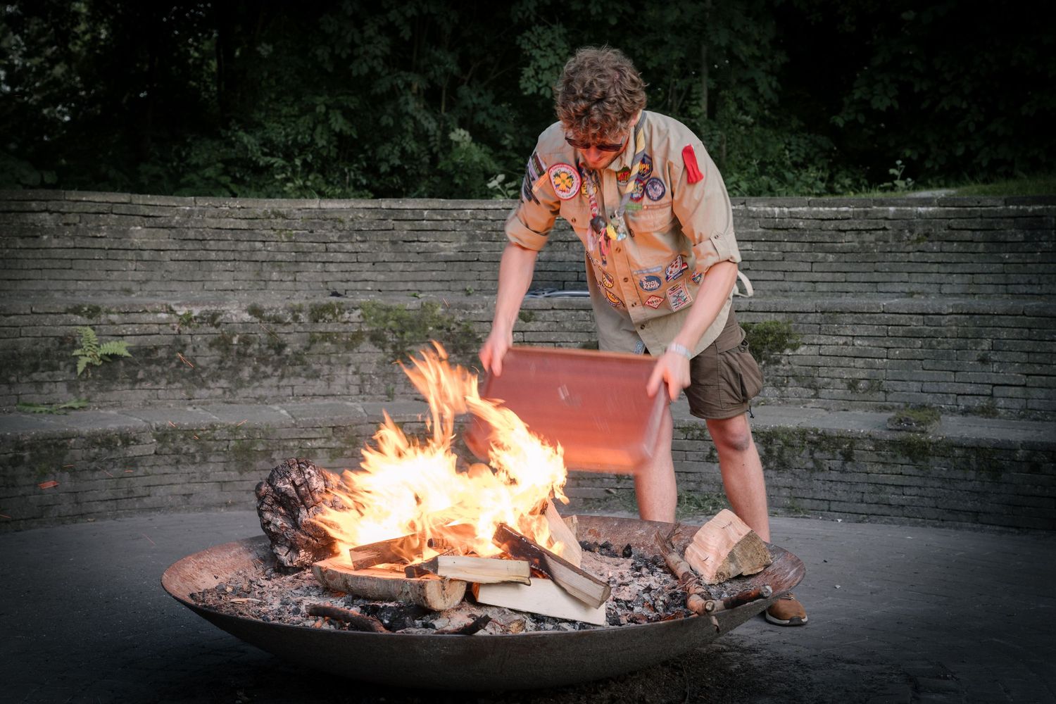 Scout leader in uniform tends to a blazing campfire in a stone fire pit at dusk.