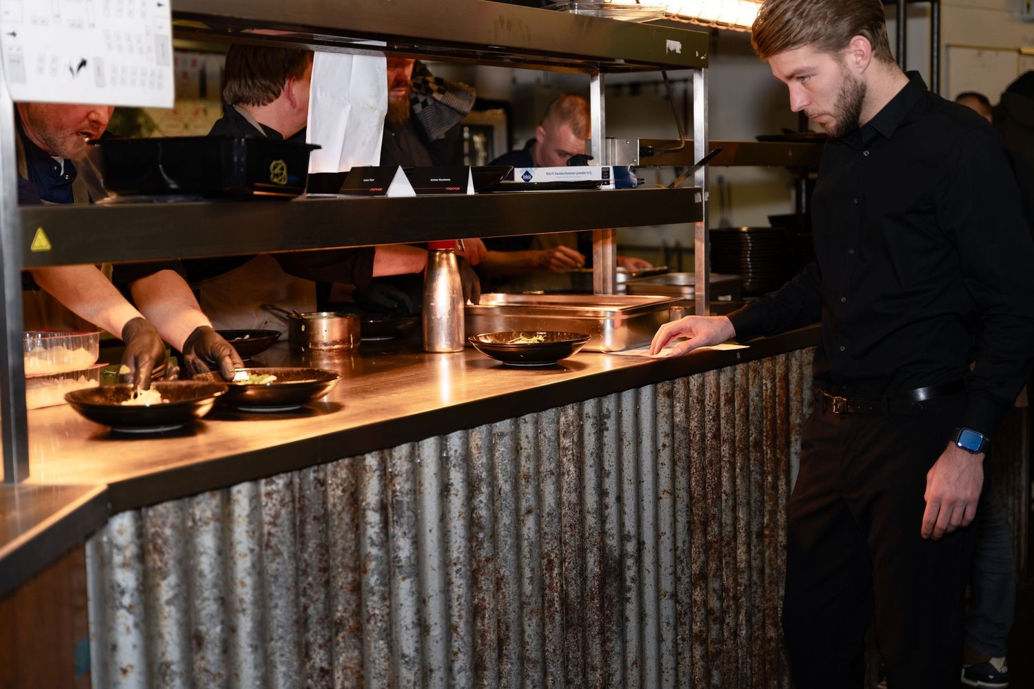 Restaurant worker stands at rustic counter with corrugated metal paneling in dimly lit kitchen service area.