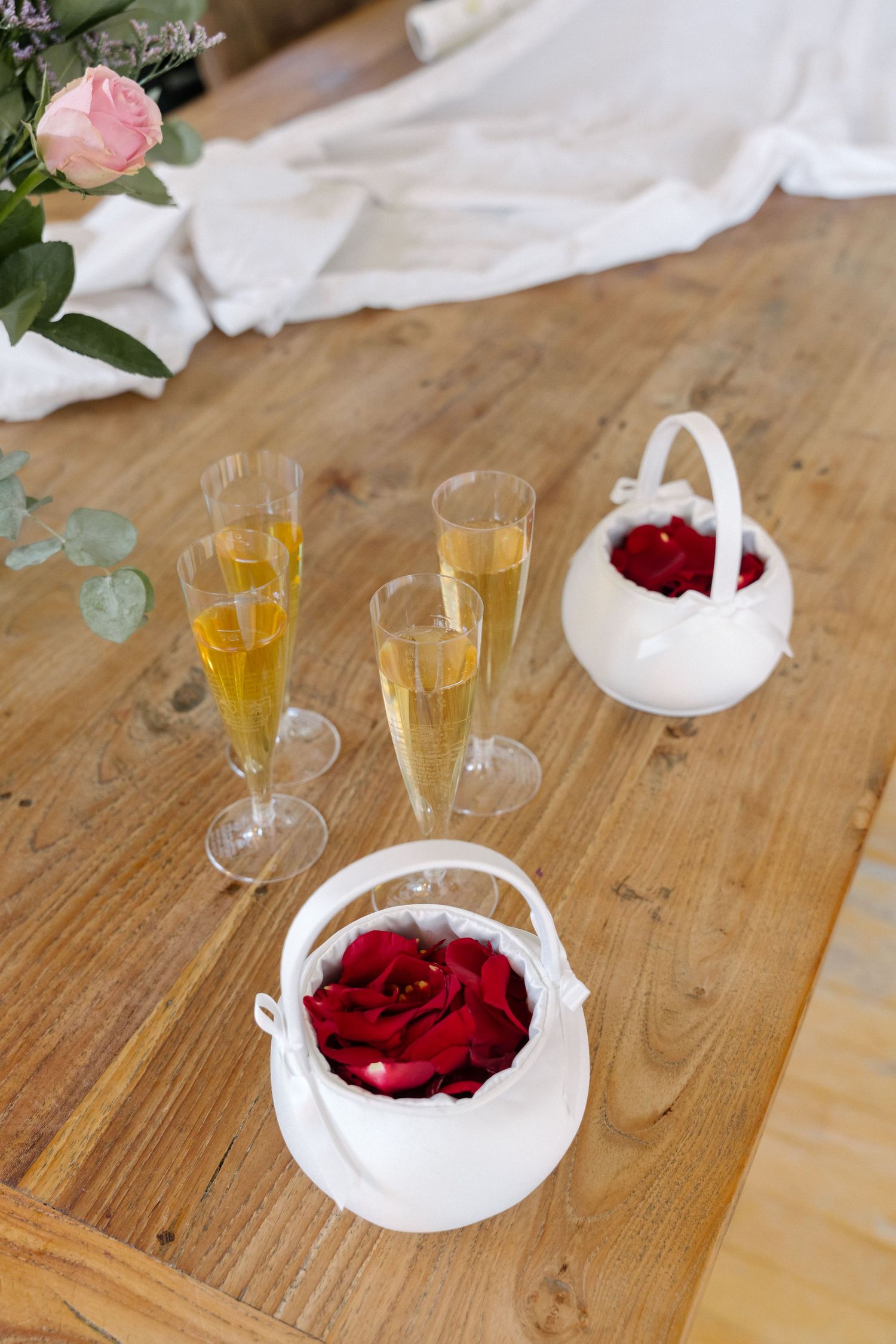 Champagne flutes and white flower baskets with red roses arranged on a rustic wooden table for a wedding celebration.