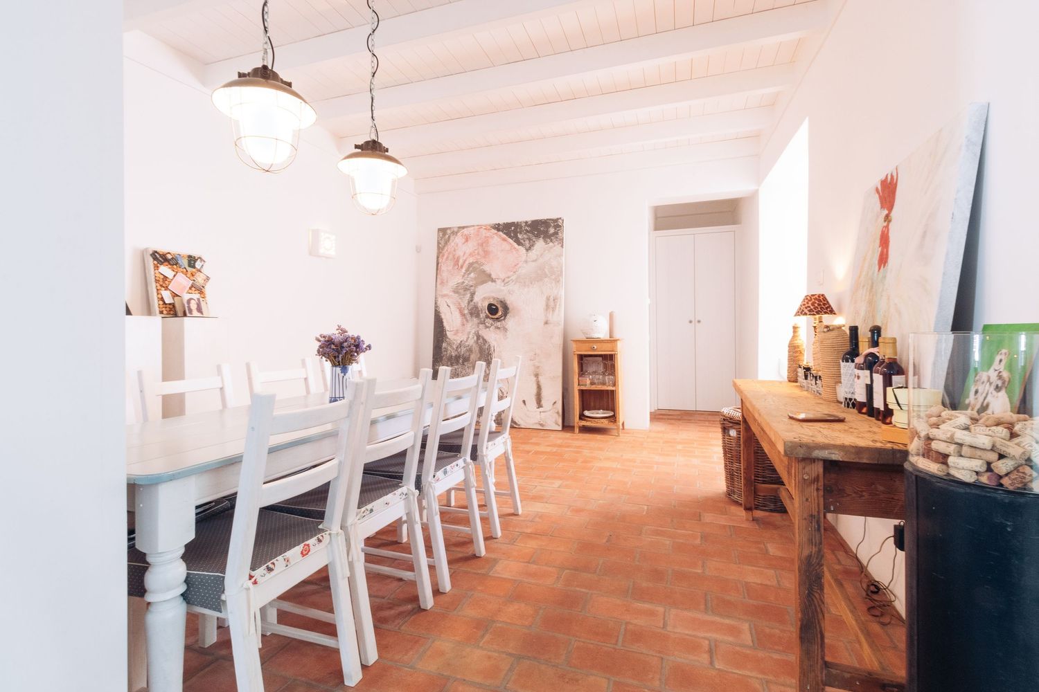 Long dining room with white chairs, pendant lights, and terracotta flooring.