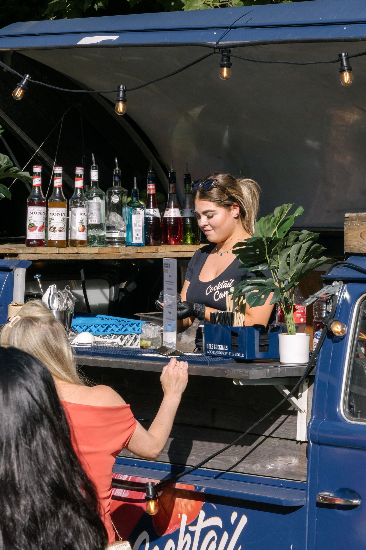 A mobile bar setup with string lights serves customers from a vintage vehicle outdoors with bottles of alcohol on display.