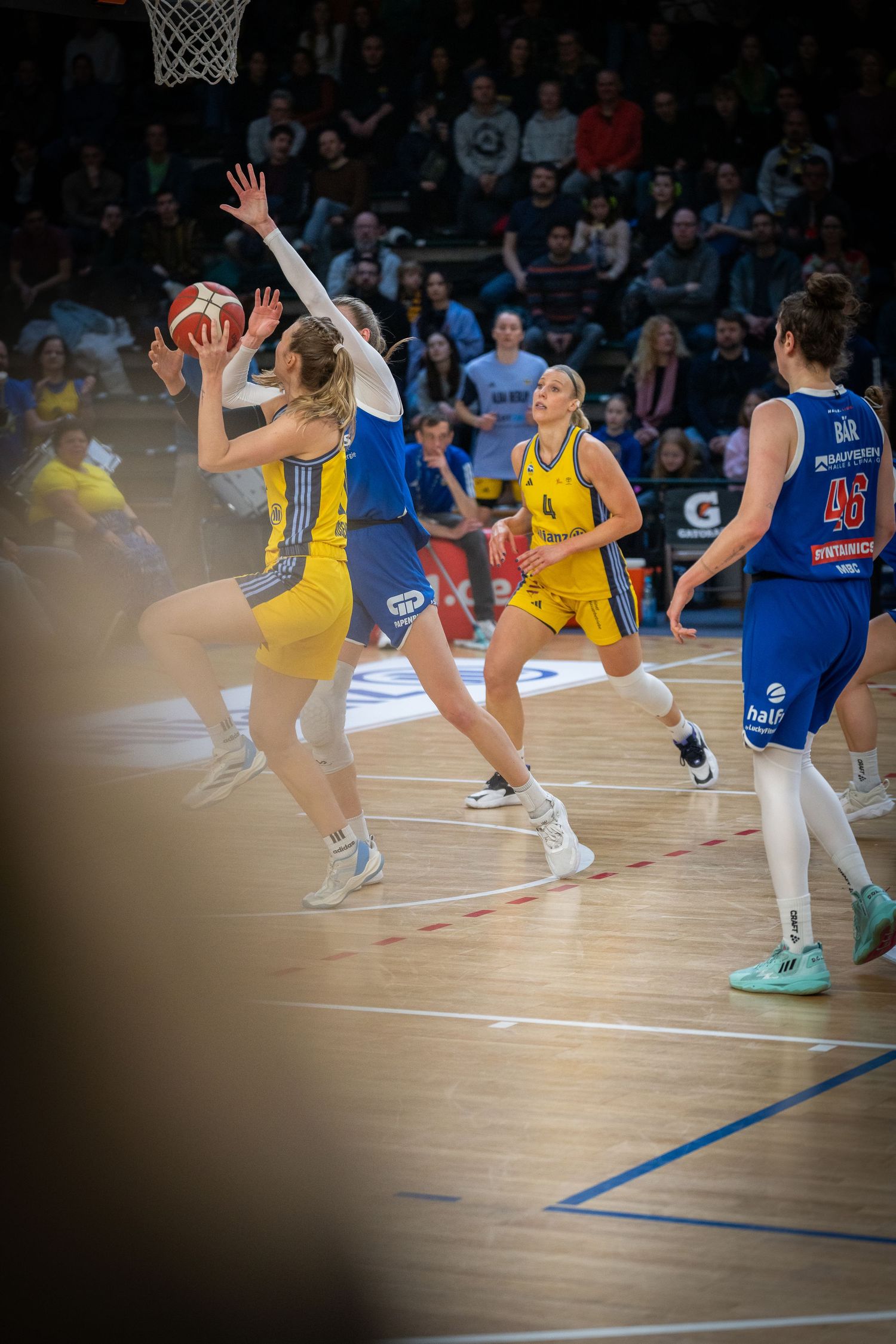 Basketball players in yellow and blue uniforms compete for possession during an intense game on the hardwood court.