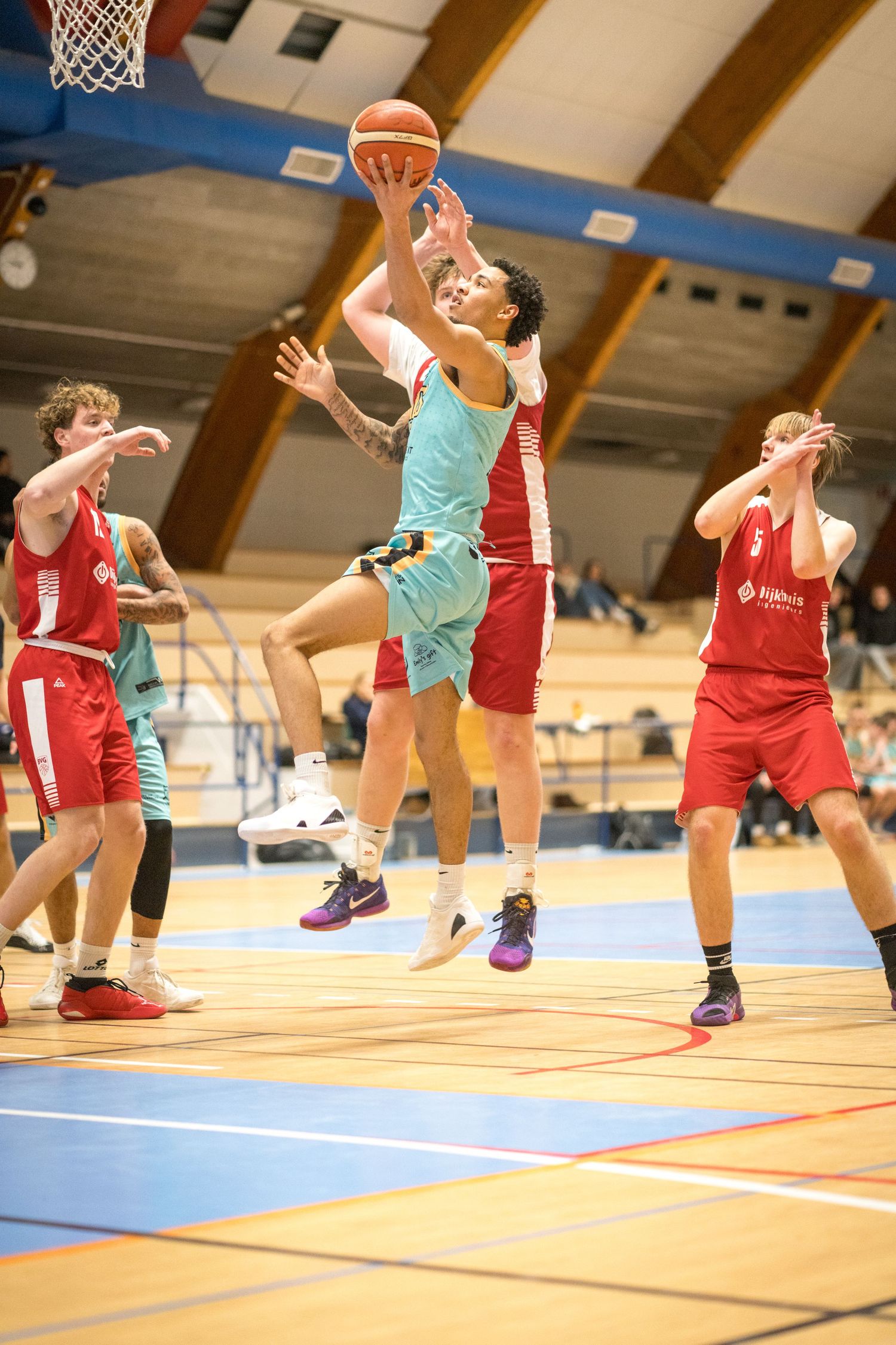 Basketball player in light blue jersey jumps for a layup between defenders in red uniforms during indoor game.