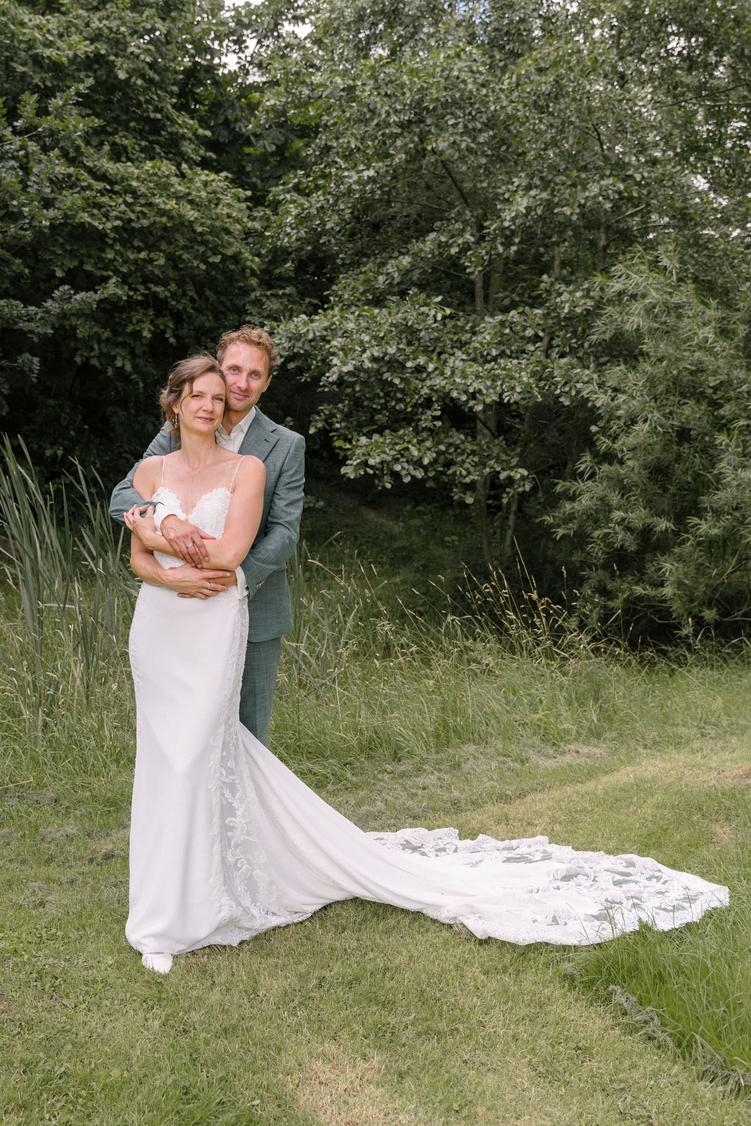 A couple poses together outdoors in formal attire with the bride wearing a flowing lace train wedding dress in a garden setting.