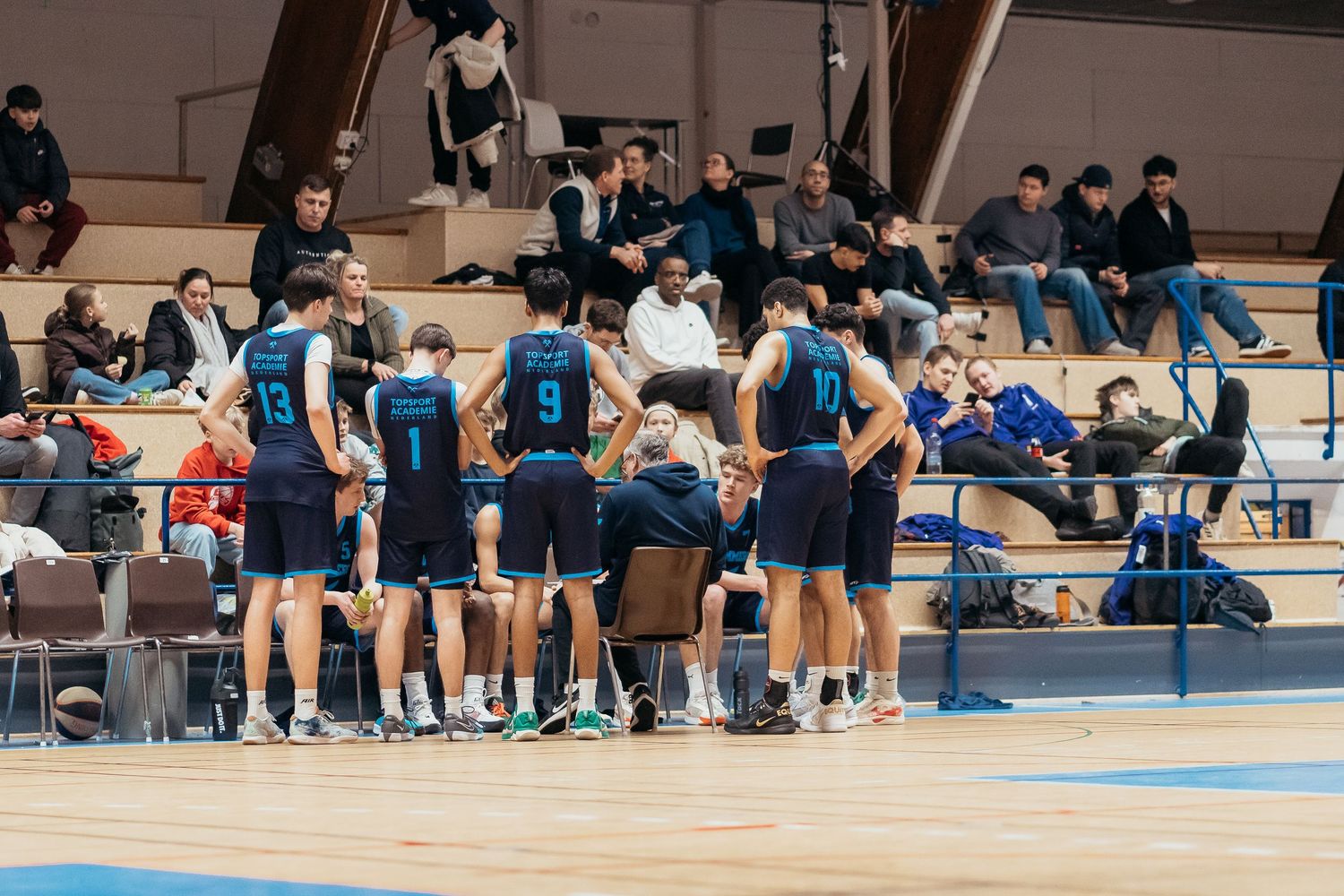 Sports team huddles on sideline during indoor volleyball game while spectators watch from bleachers in gymnasium.