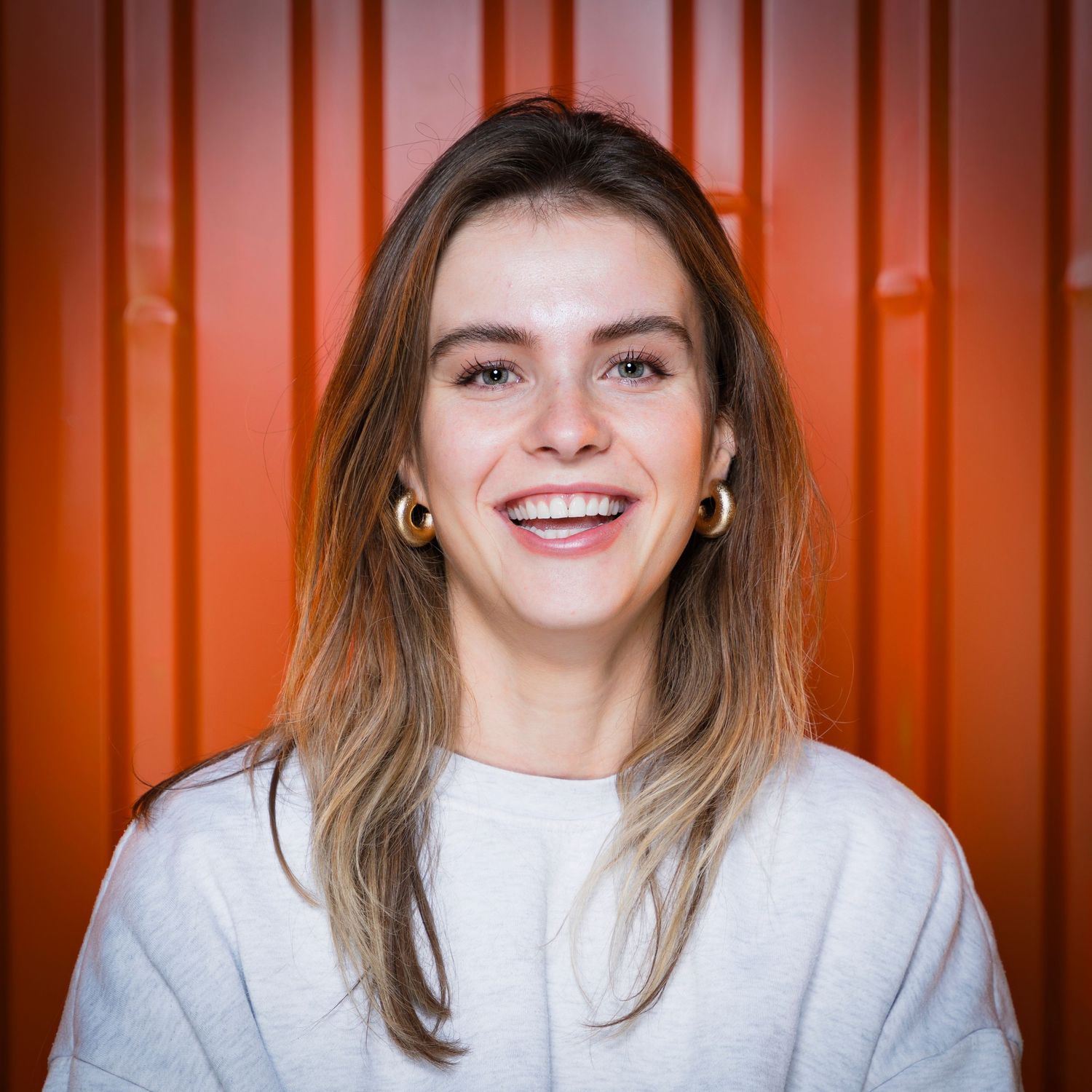 Woman wearing white sweater smiles warmly against orange corrugated metal wall background.