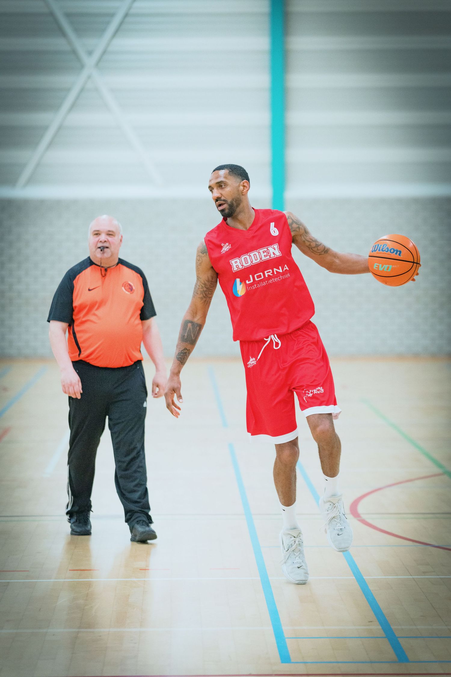 A basketball player in red dribbles near the sideline while an official in orange observes the play.