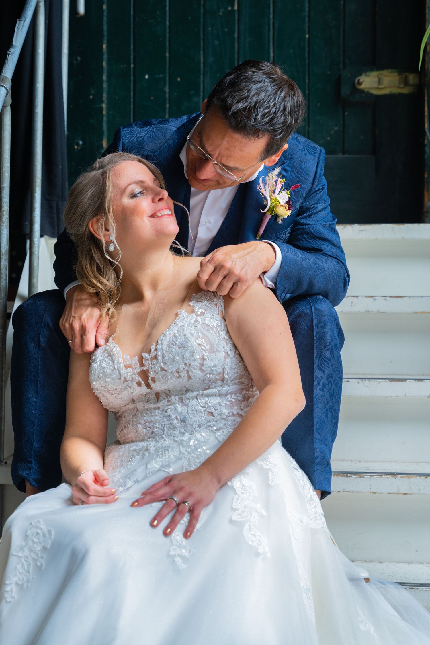 Couple poses intimately on white steps, with groom in blue suit leaning over bride in lace wedding dress.