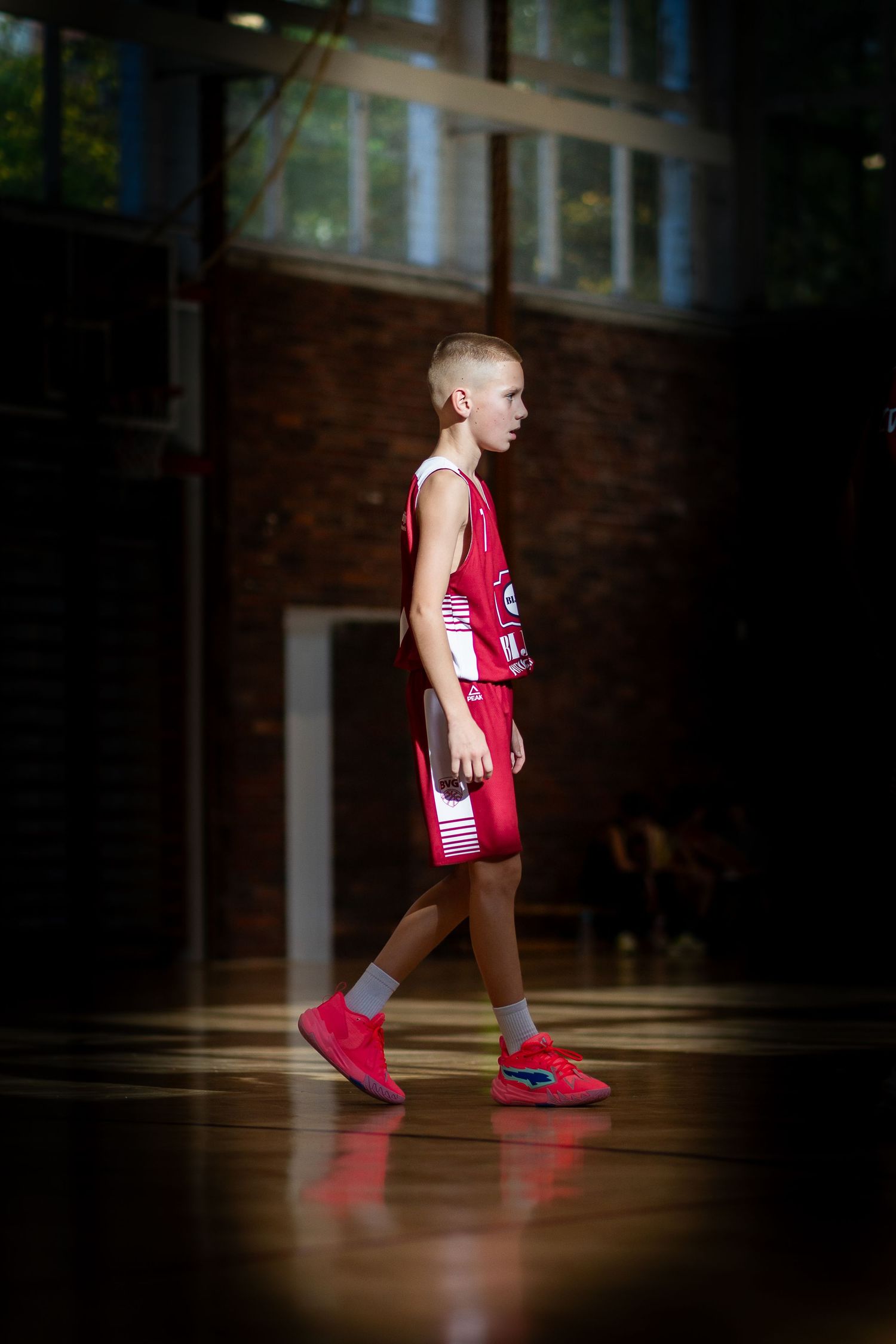 A basketball player in a red jersey and matching shoes walks across a polished gymnasium floor in dramatic lighting.