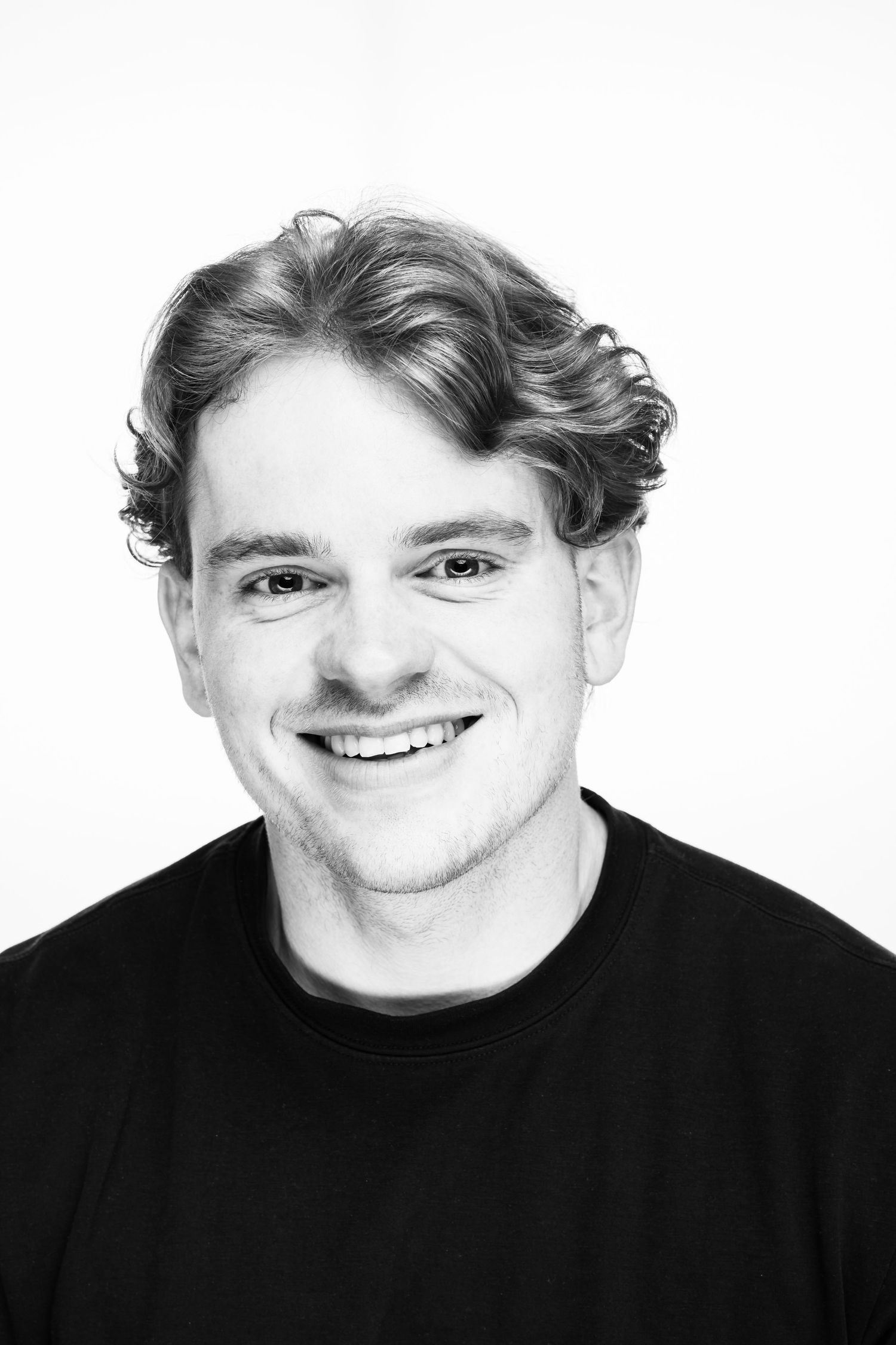 Black and white headshot portrait of a person with wavy hair wearing a black t-shirt and smiling at the camera.