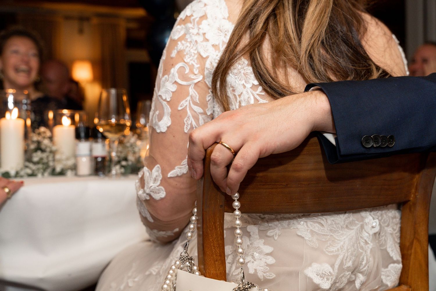 Bride in a white lace wedding dress with sheer sleeves holds hands with someone while seated at a candlelit reception table.