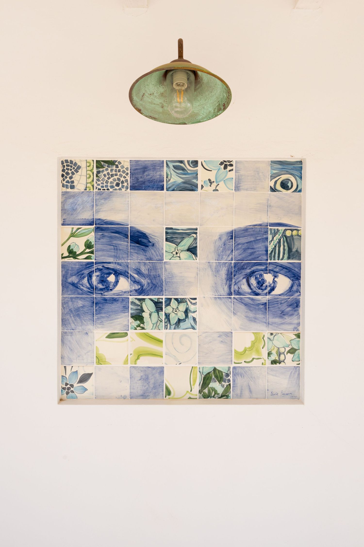Decorative blue and green ceramic tile mosaic of eyes mounted on white wall beneath copper pendant lamp.