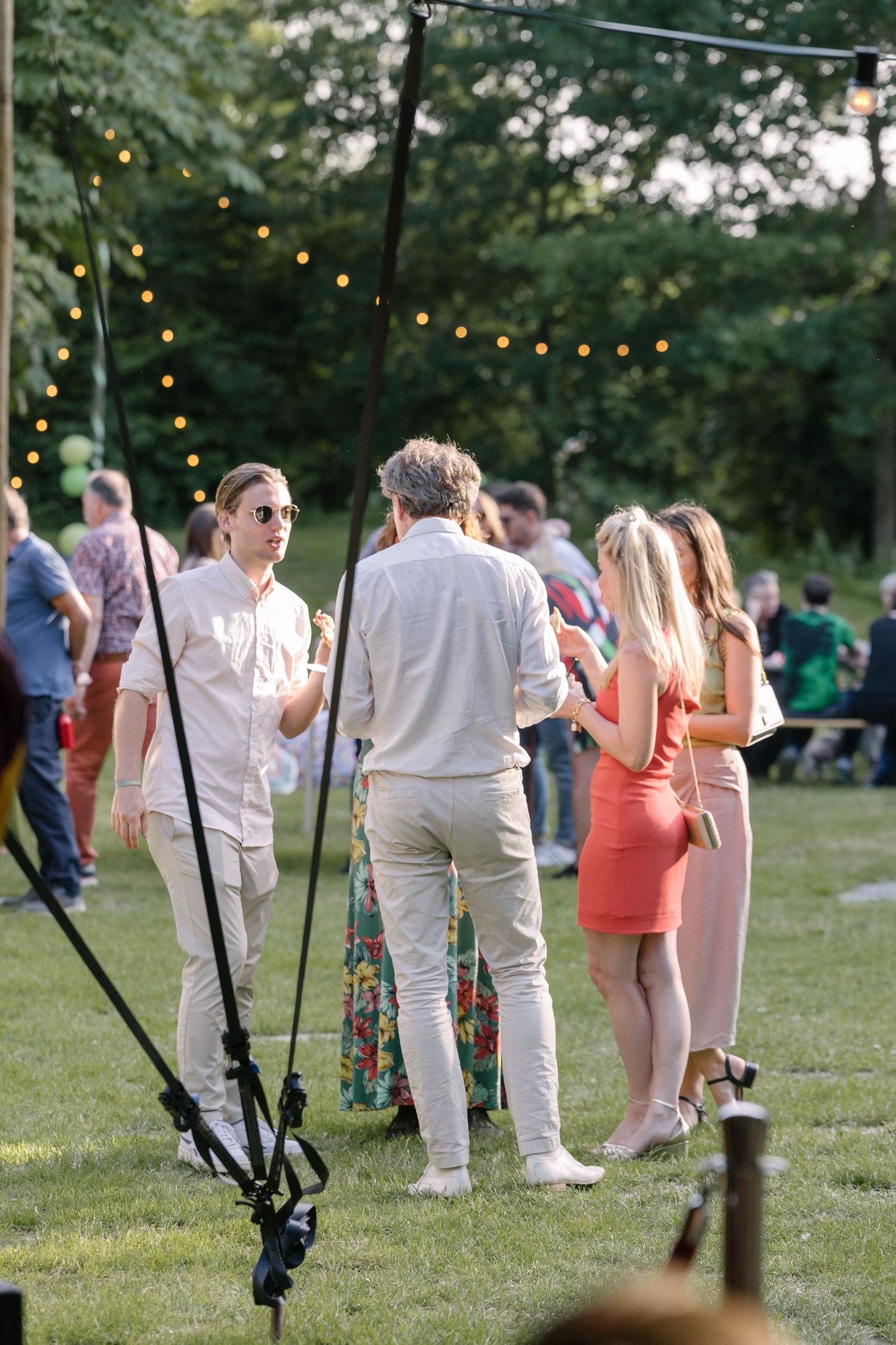 Outdoor garden party with string lights overhead as people gather on a green lawn during sunset.