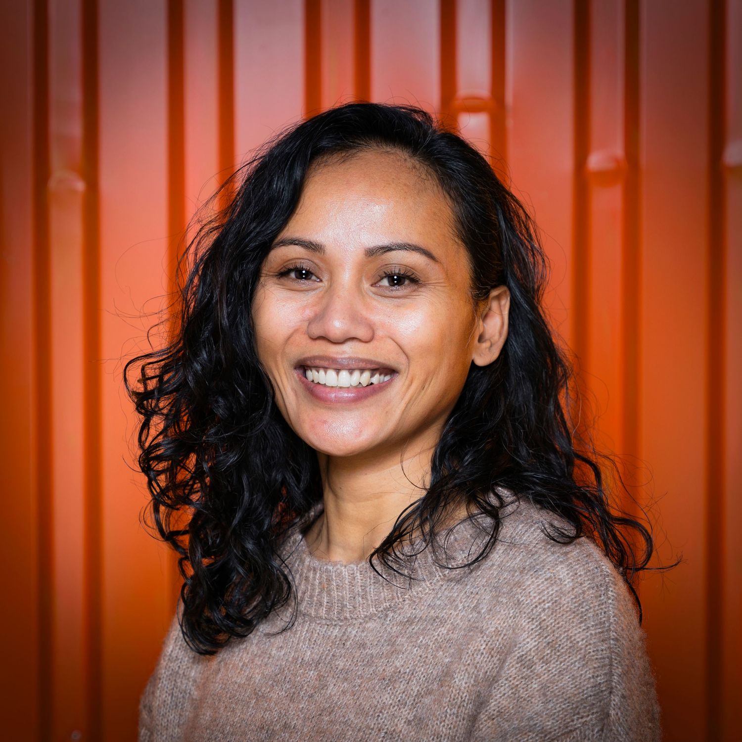 A smiling person with curly dark hair wearing a beige sweater stands in front of an orange corrugated wall.