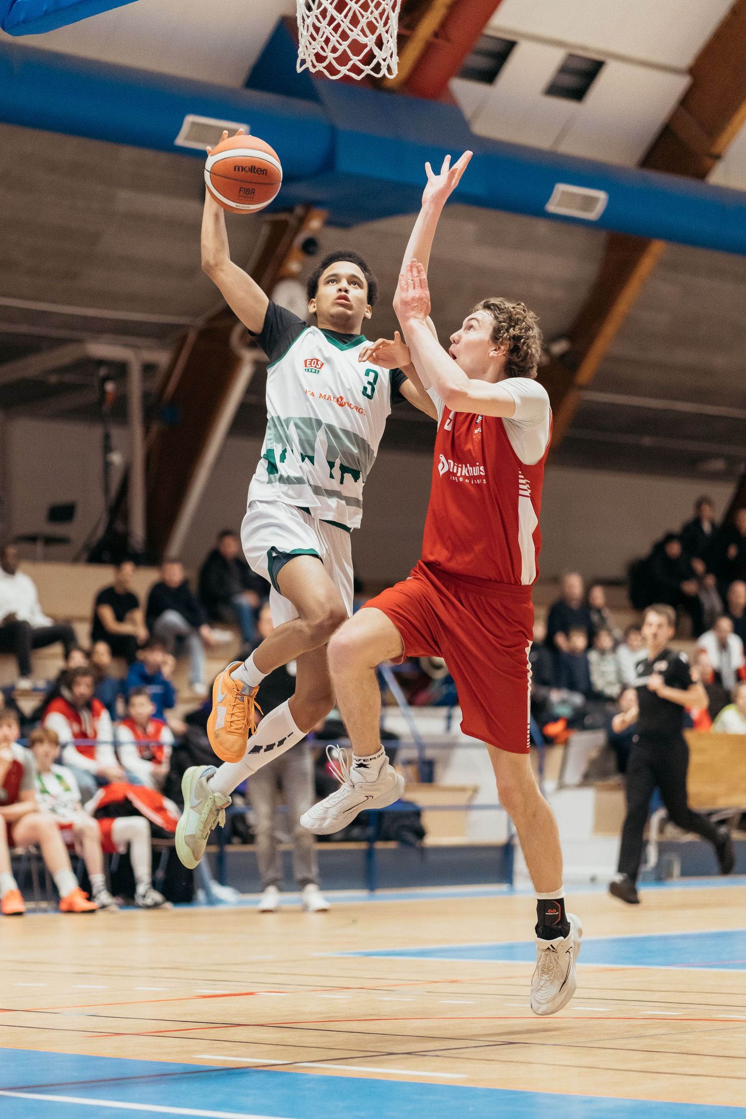 Two basketball players leap high in an indoor game, one in white attempting a shot while another in red defends.