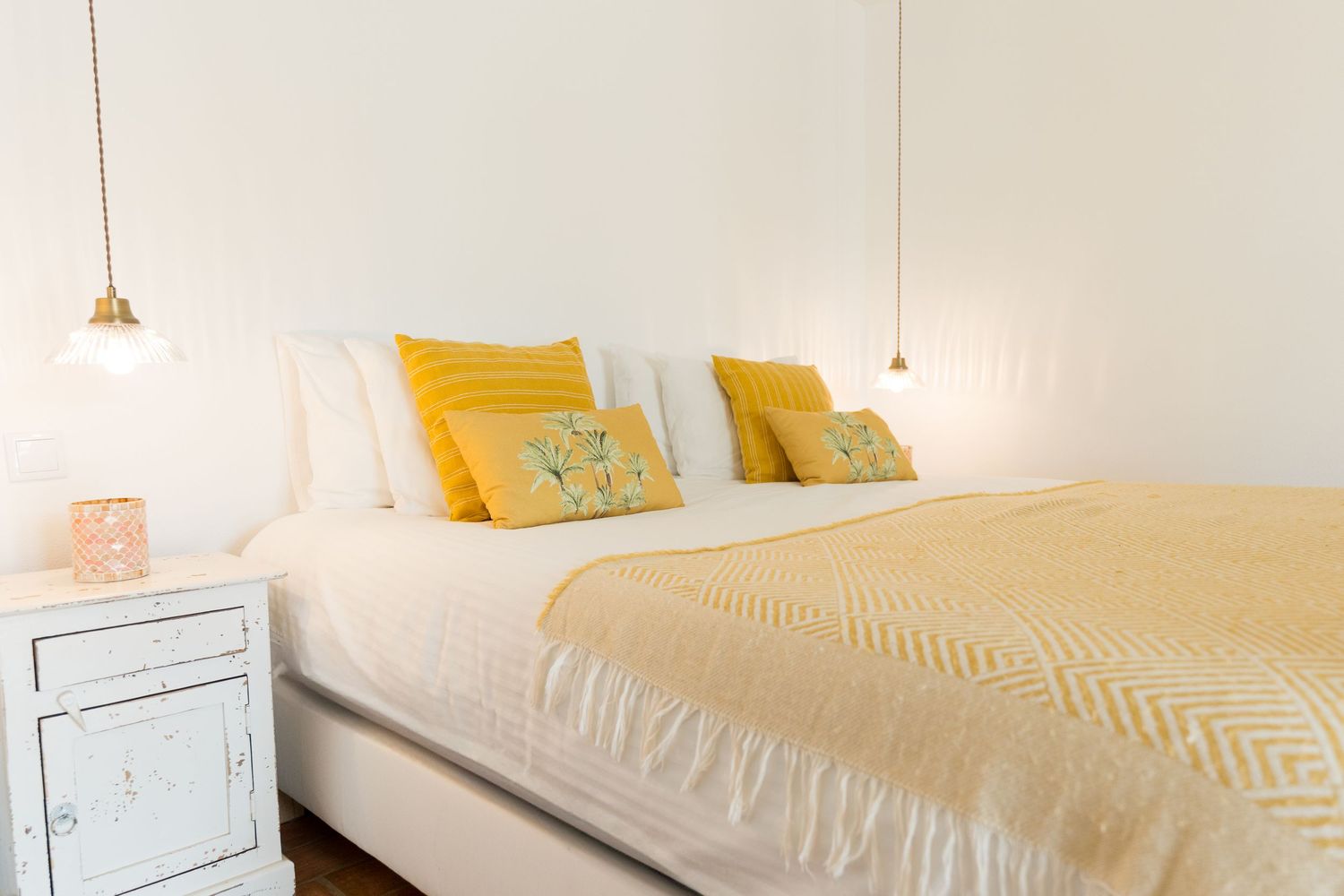Serene bedroom with white bedding, yellow accent pillows, and vintage bedside table.