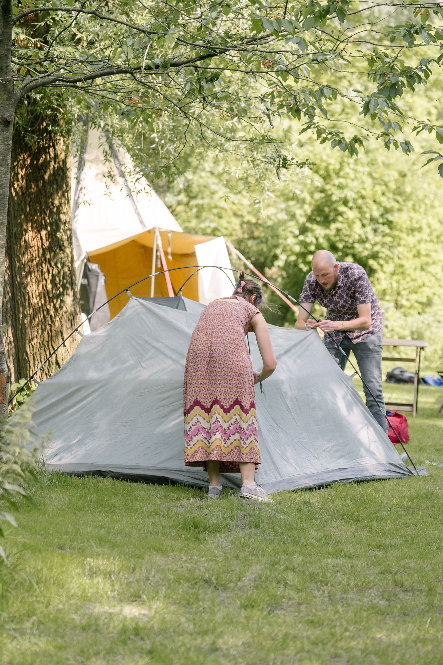 People setting up a modern gray camping tent under trees at a campsite during a sunny summer day.