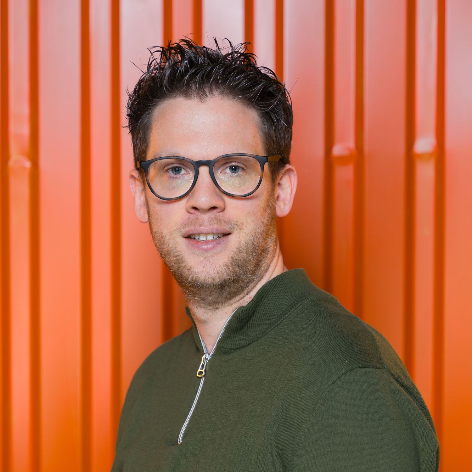A person with glasses wearing a green zip-up sweater smiles in front of an orange metal backdrop.