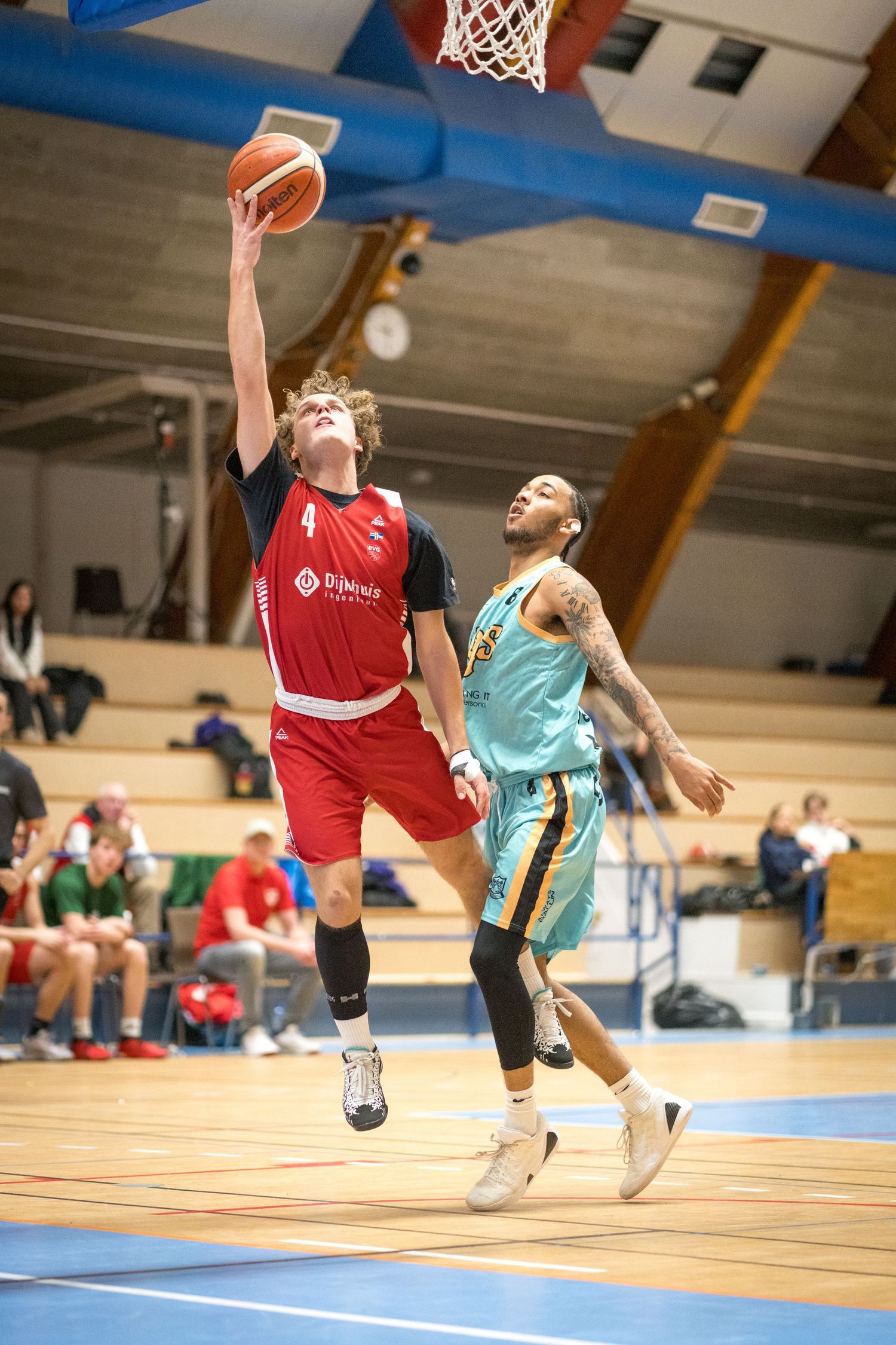 Basketball players competing in a game as one athlete in red goes for a layup against a defender in light blue.