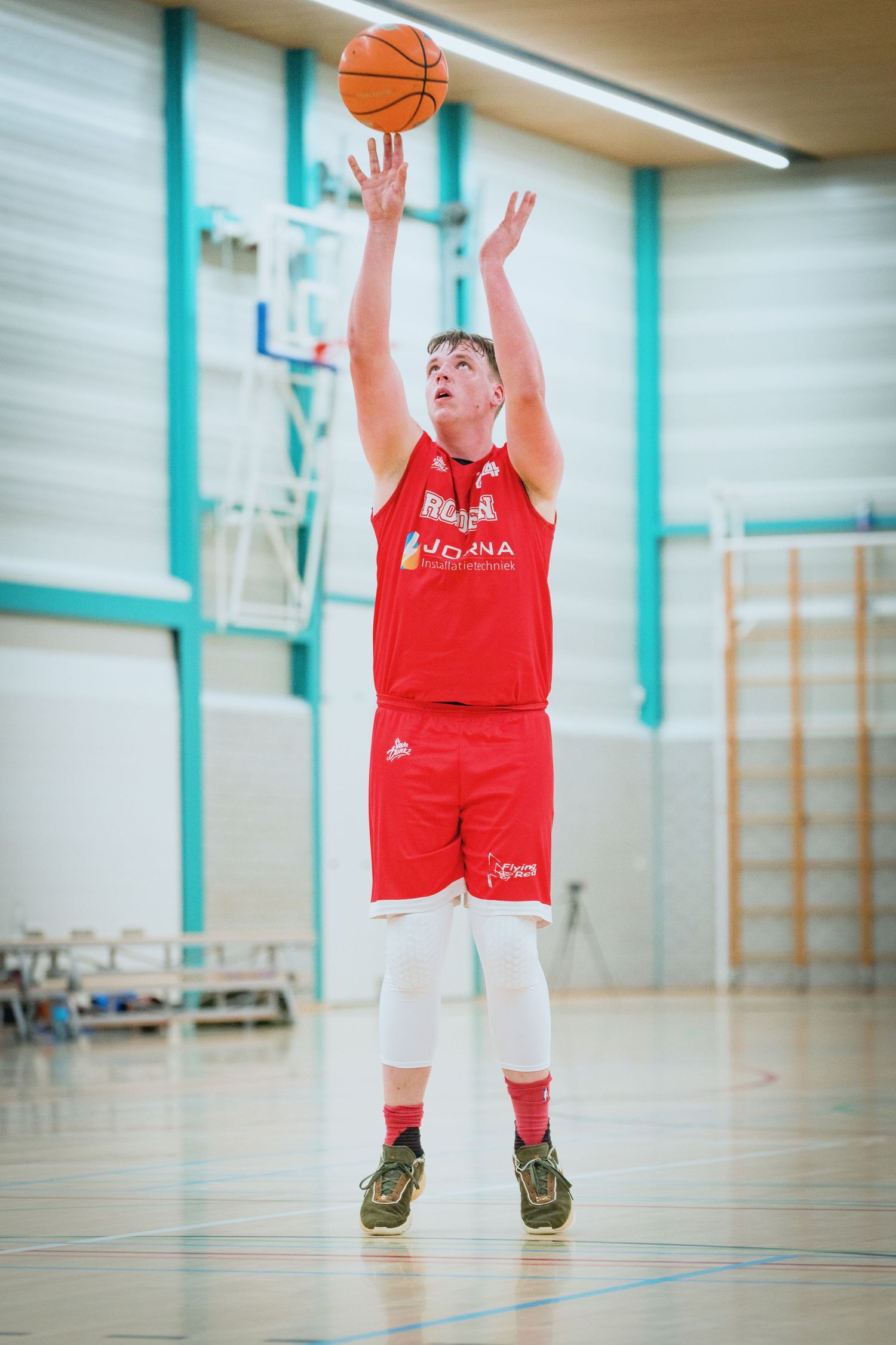 A basketball player in a red jersey takes a jump shot during practice in a gymnasium.