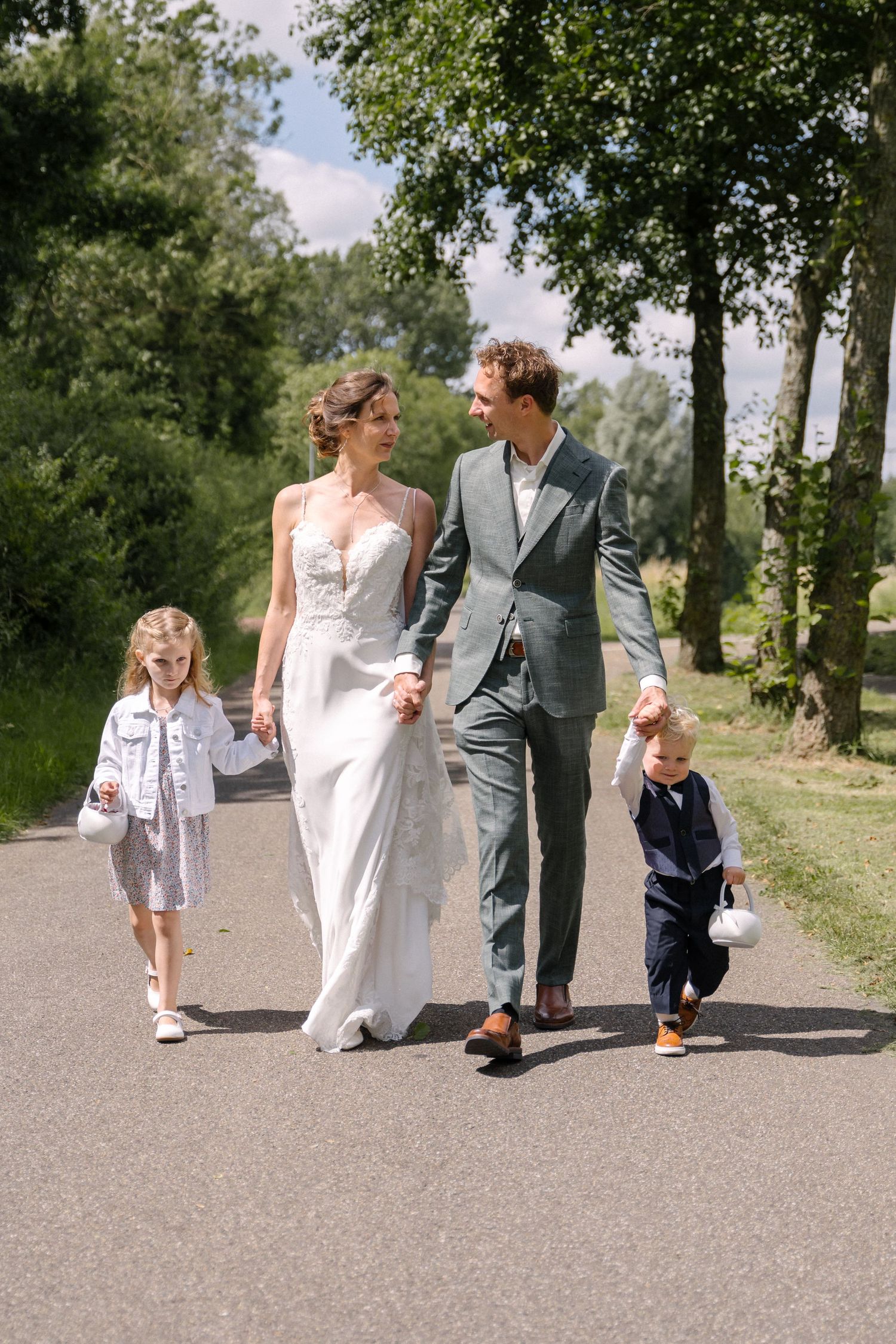 Wedding couple walking together on path with two small children, surrounded by green trees on sunny day.