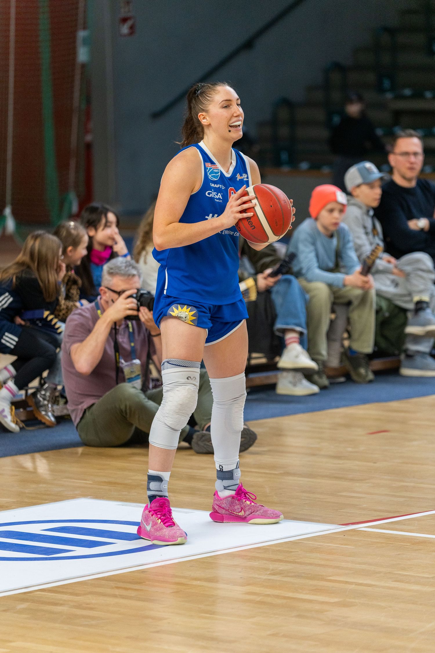 Basketball player in blue uniform with white knee pads and pink shoes holds ball during indoor game with spectators.