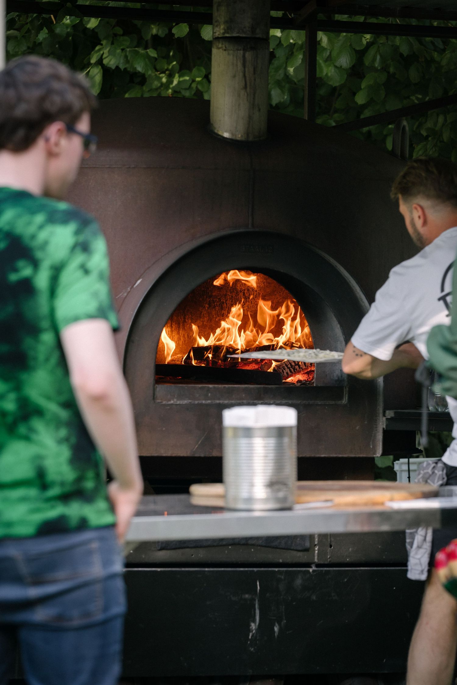 Wood-fired pizza oven with roaring flames being tended to in an outdoor kitchen setting.