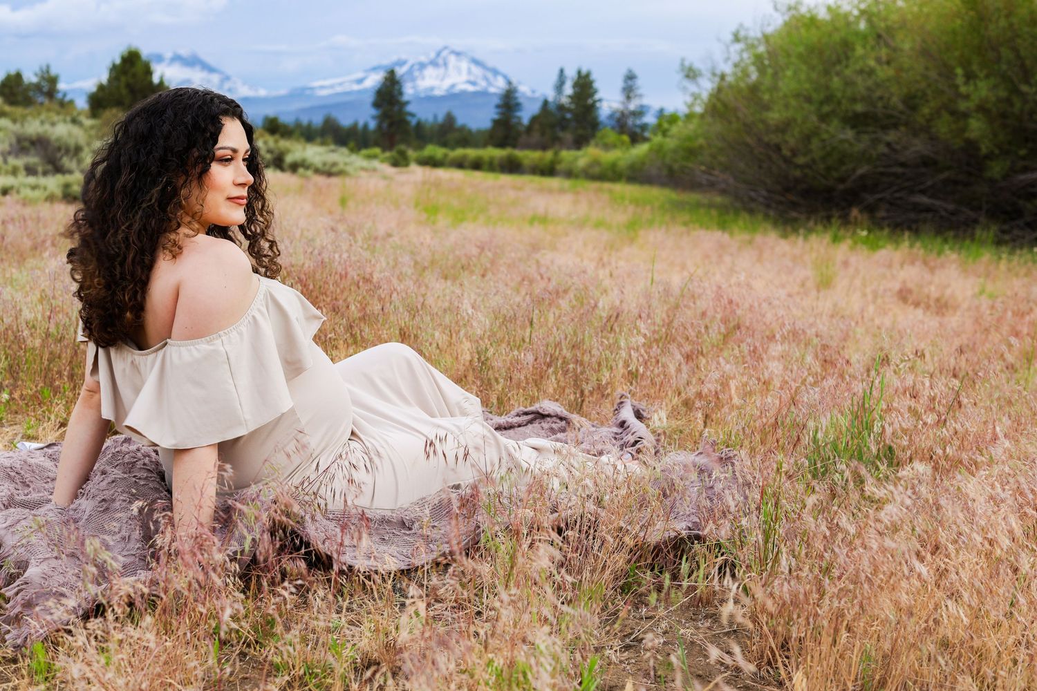 A person in a flowing white dress sits gracefully in a golden meadow with snow-capped mountains visible in the distance.