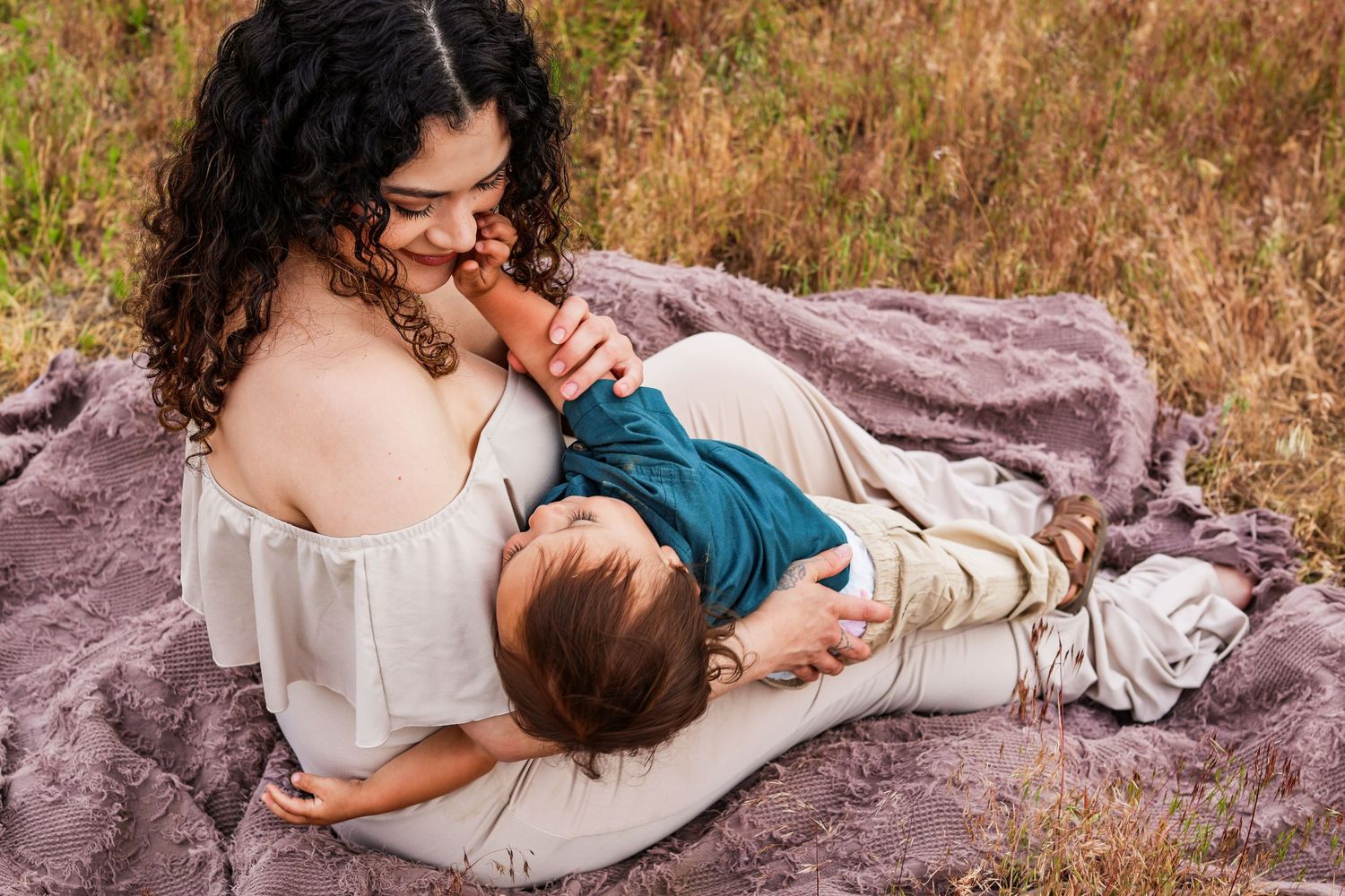Mother and baby share tender moment while resting on purple blanket in autumn field during outdoor photoshoot.