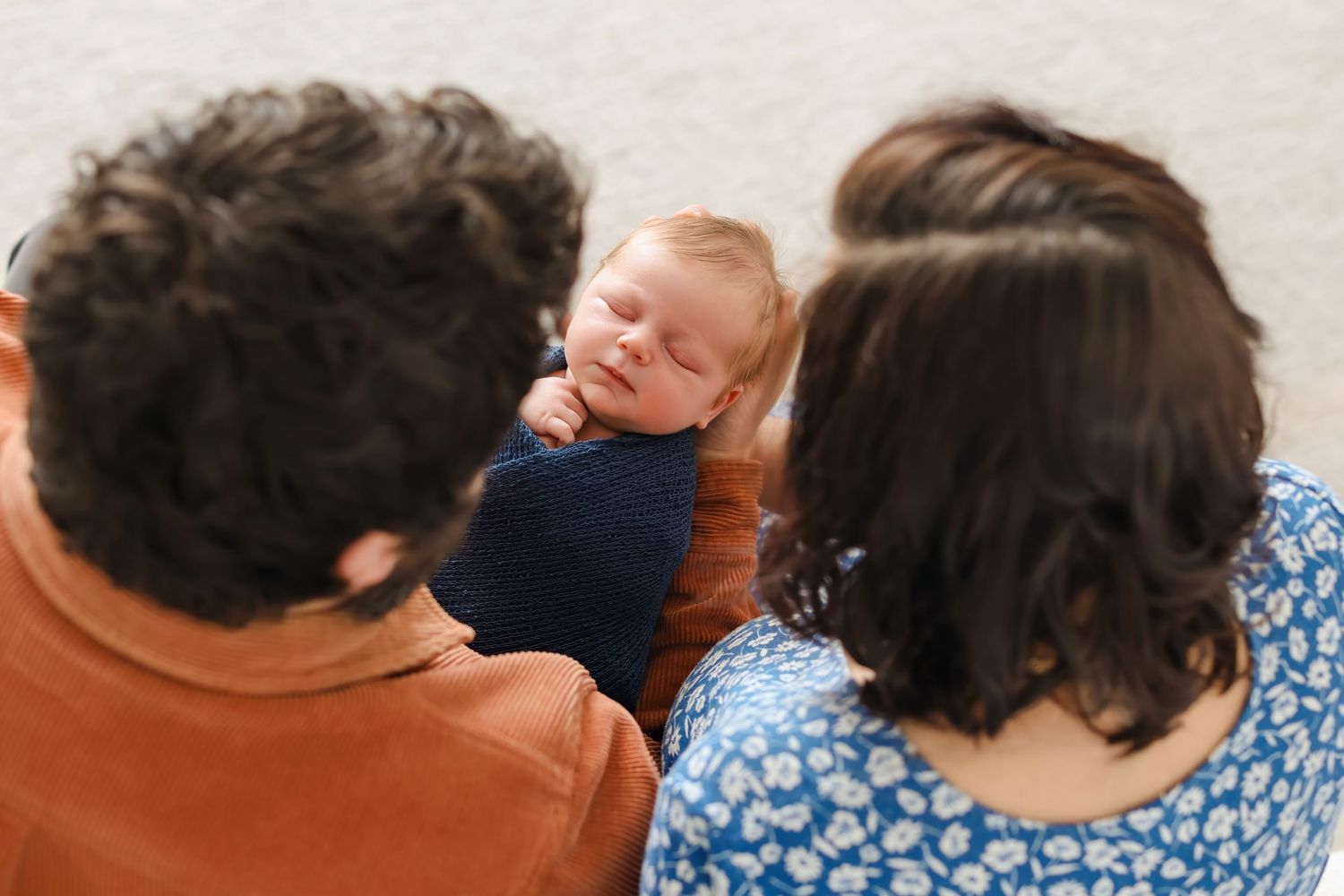A sleeping baby in a navy onesie being held while resting between two adults wearing colorful shirts.