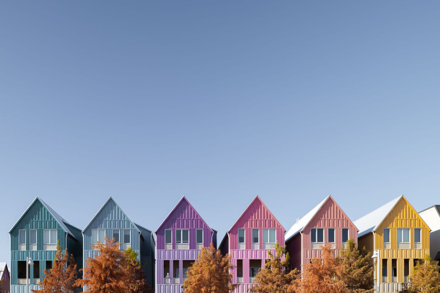 Colorful row houses in teal, purple, pink, coral and yellow stand against a bright blue sky with autumn trees below.