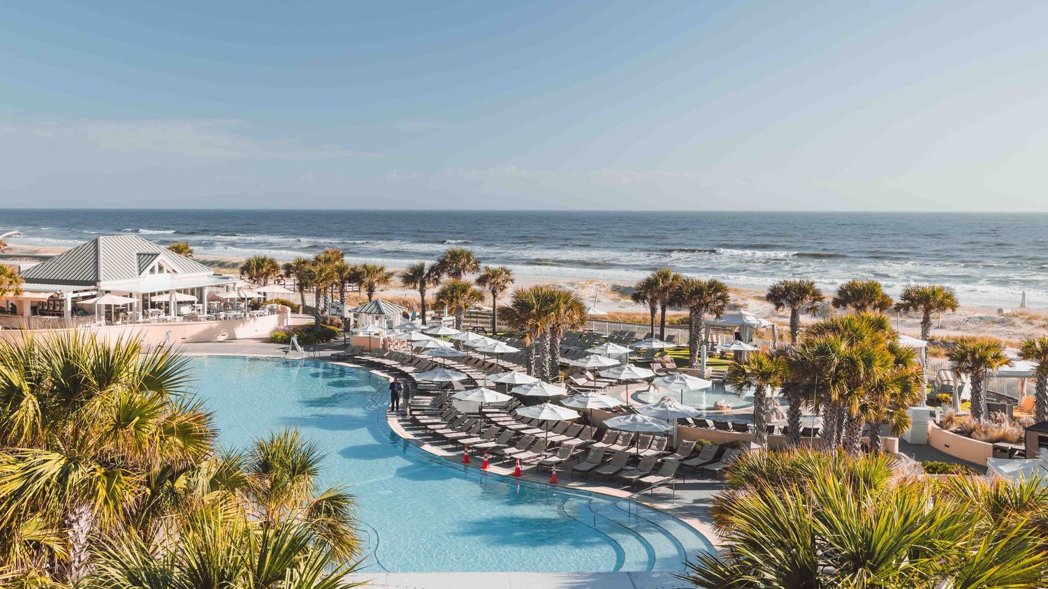 Luxurious beachfront resort with winding pool, palm trees and lounge chairs overlooking the ocean on a sunny day.