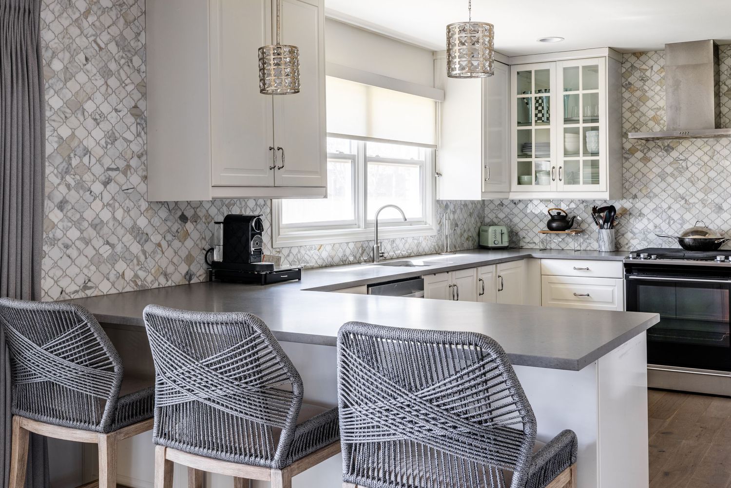 Modern white kitchen with gray counter stools, quartz countertops and geometric tile backsplash against white cabinets.