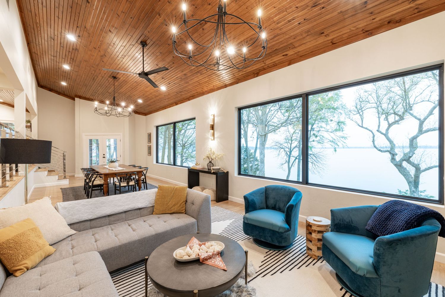 Modern open concept living room with wood ceiling, large windows, gray sofa and teal accent chairs in coastal style decor.