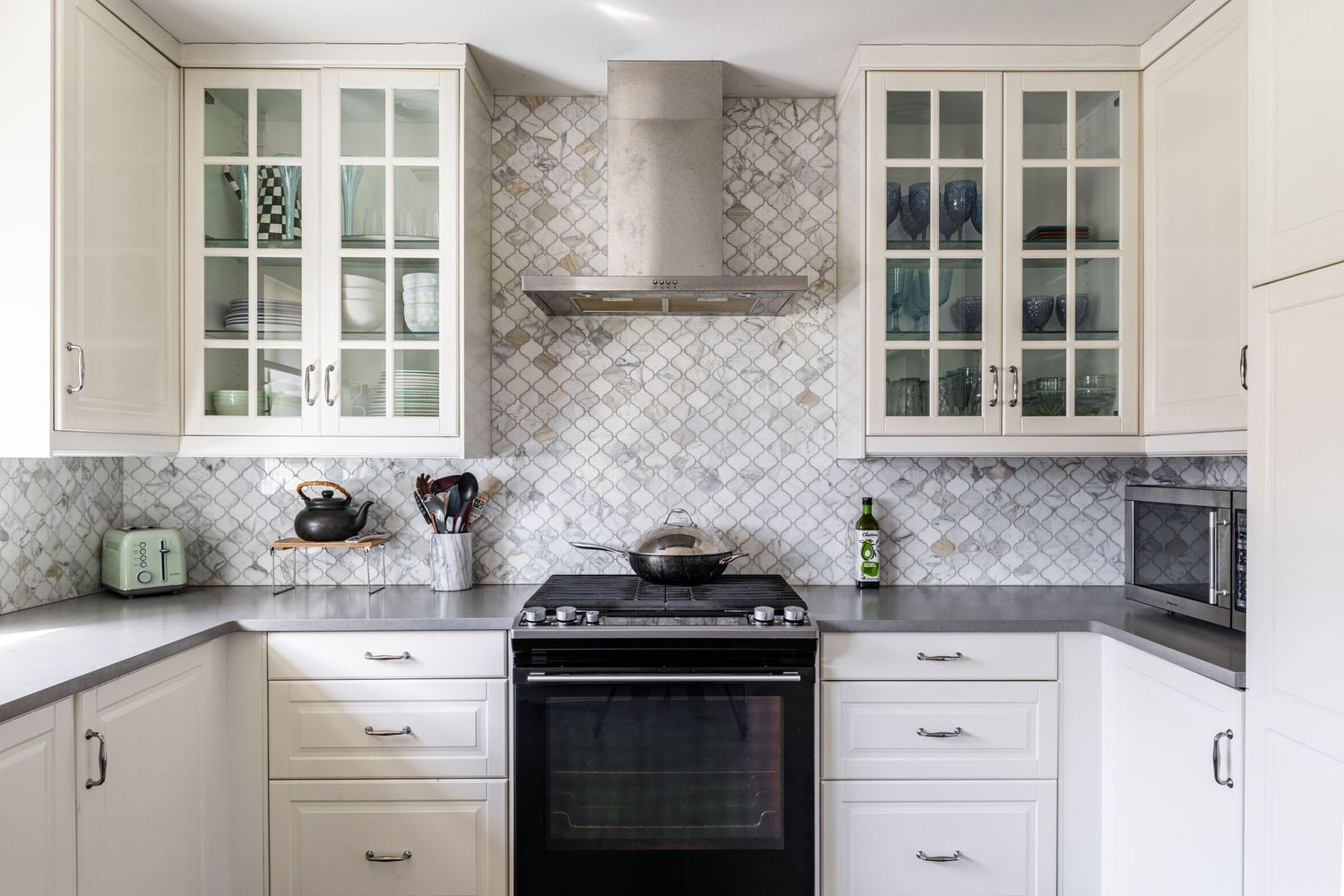 Modern white kitchen with glass-front cabinets, gray herringbone tile backsplash, and black stainless steel appliances.
