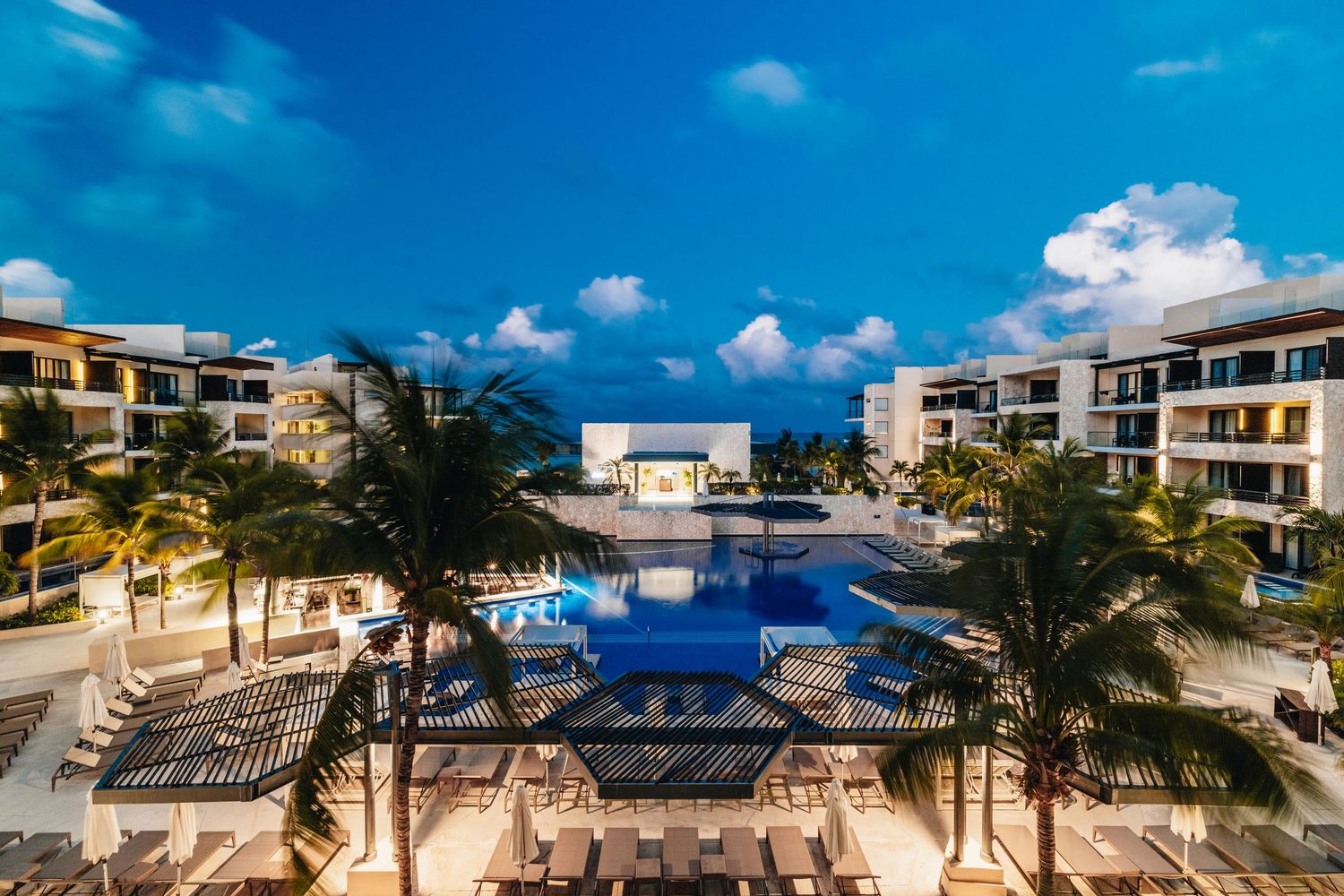 Luxury beachfront resort with swimming pool and palm trees under bright blue sky and white clouds.