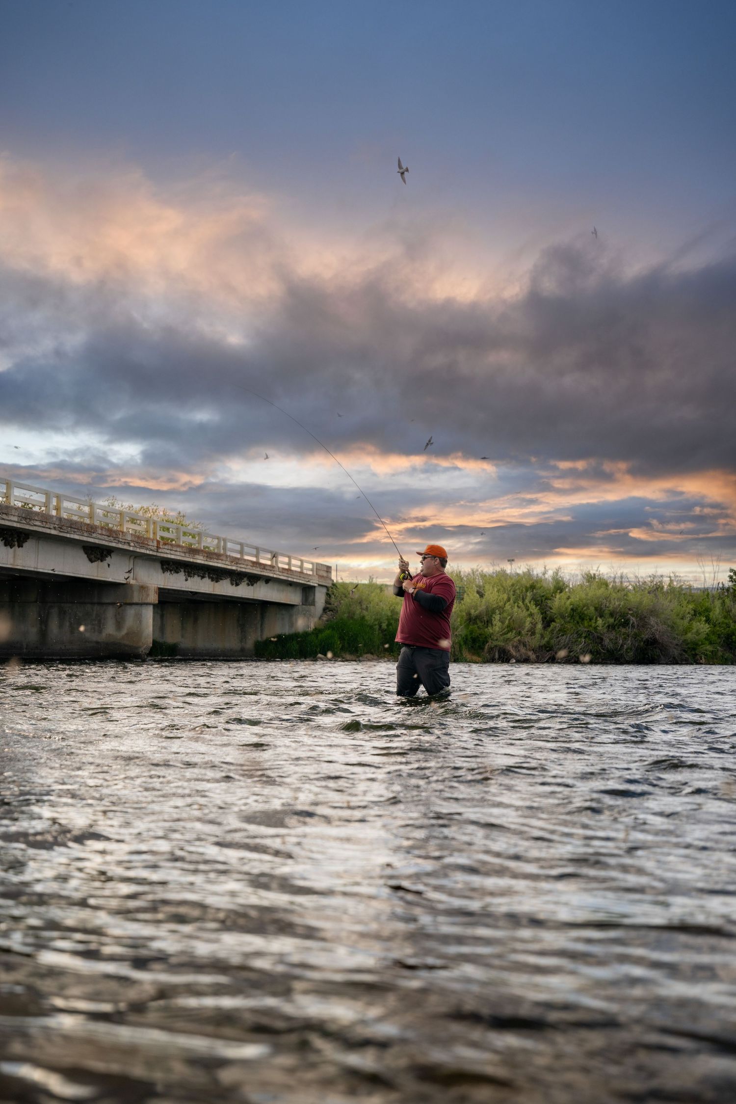 Low-angle view from water level of fisherman underneath a bridge during dramatic sunset with cloudy skies.