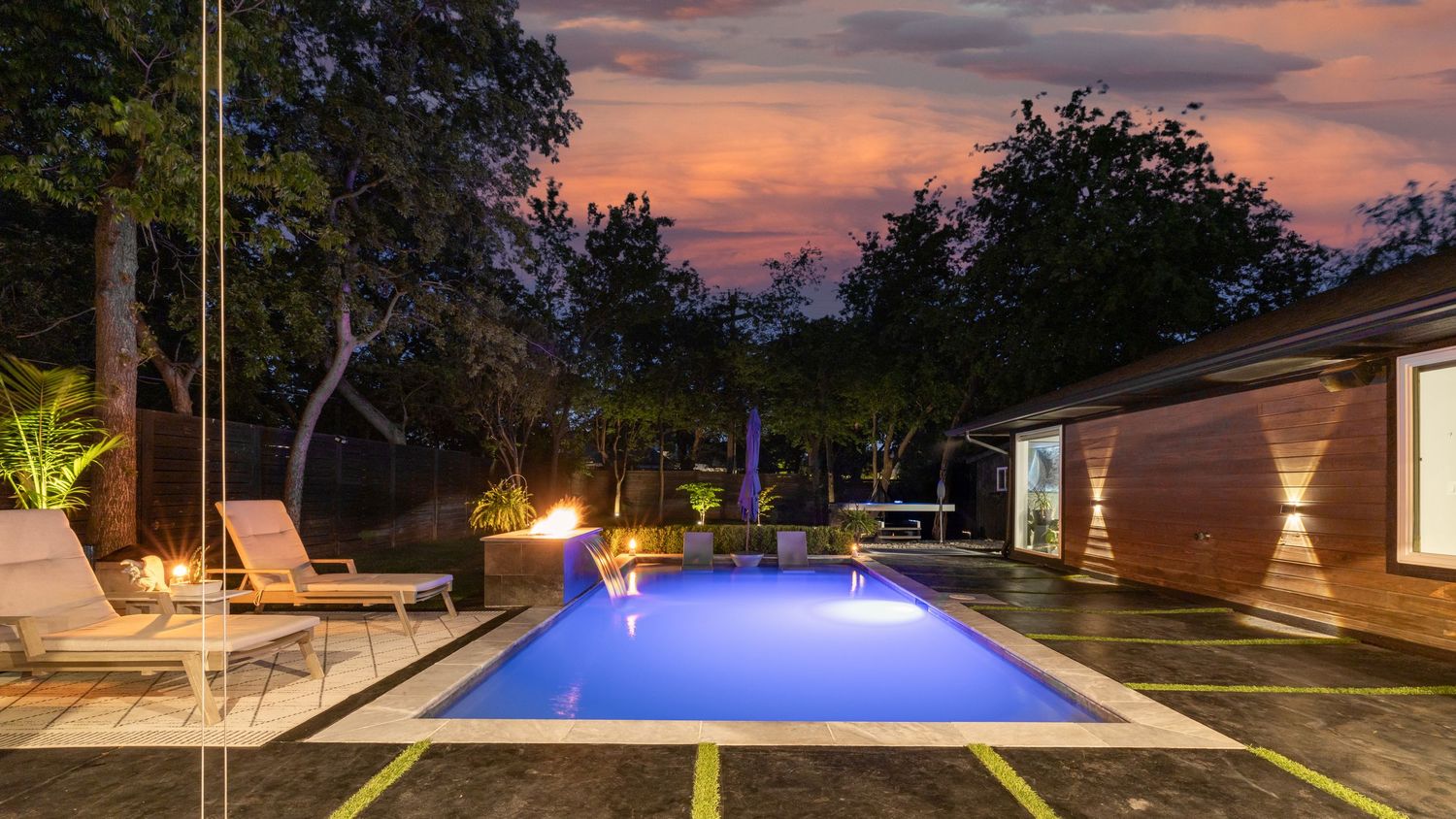 Modern backyard pool illuminated in blue with lounge chairs and landscape lighting at sunset creates a serene evening ambiance.