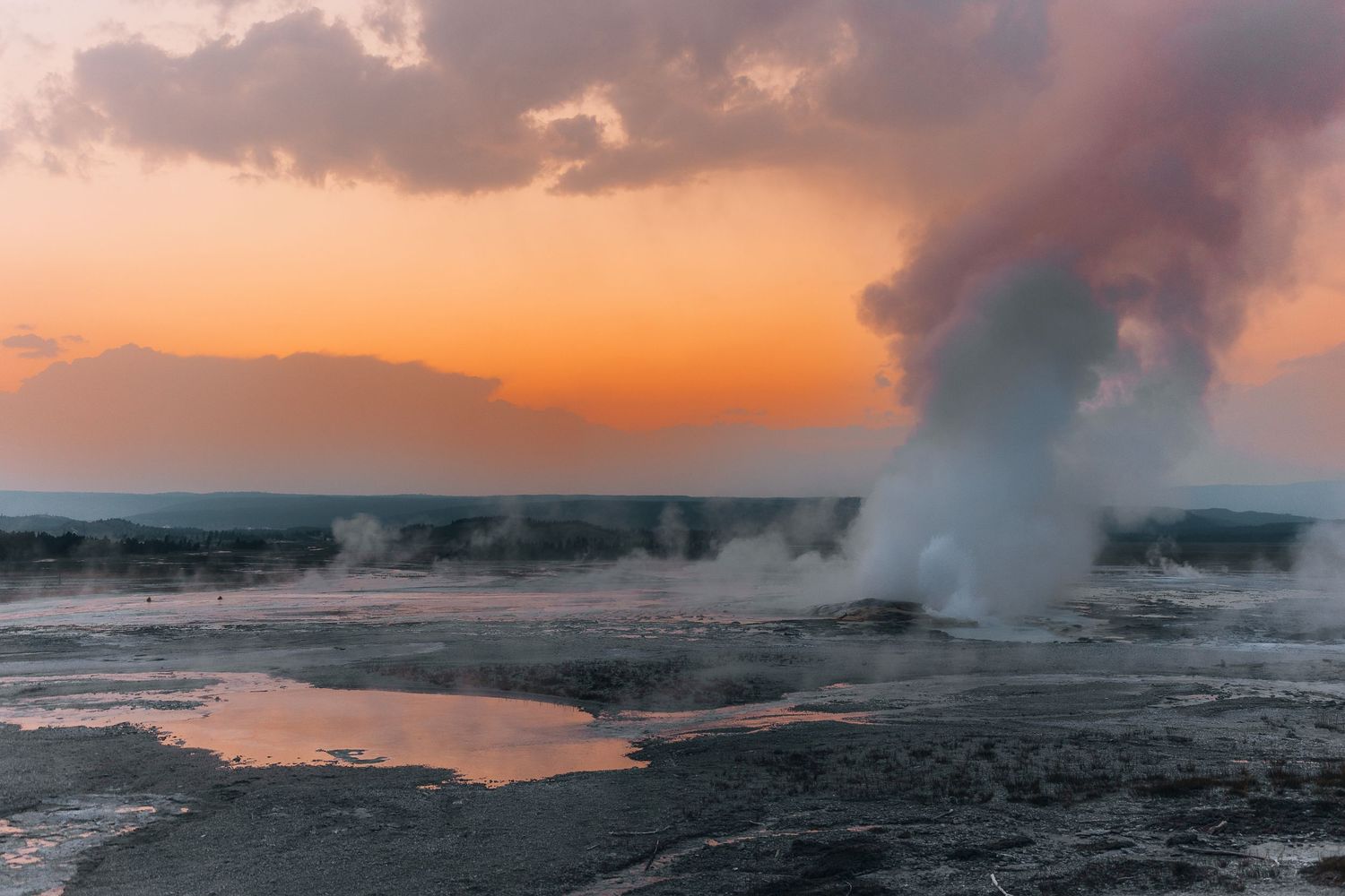 Steam rises from a geothermal geyser pool at sunset with orange sky and clouds in Yellowstone National Park.