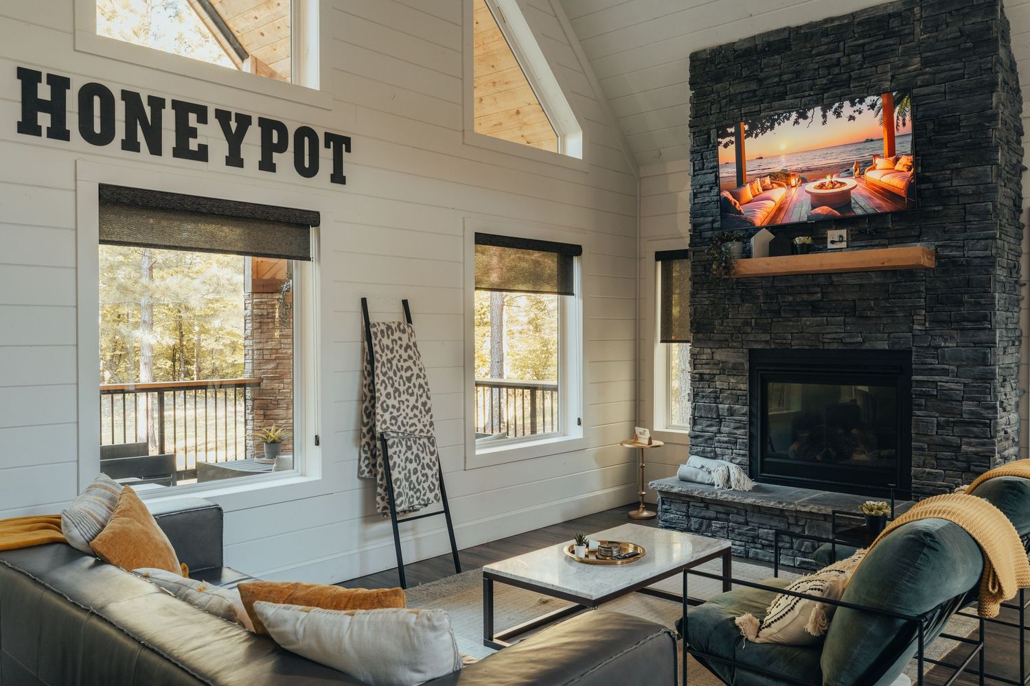 Modern living room with dark stone fireplace, honeypot sign, large windows, and gray sectional sofa in a vaulted ceiling space.