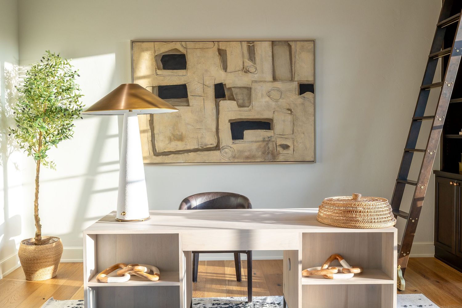 Modern home office space with white desk, potted tree plant, brass lamp and abstract black and white artwork on wall.
