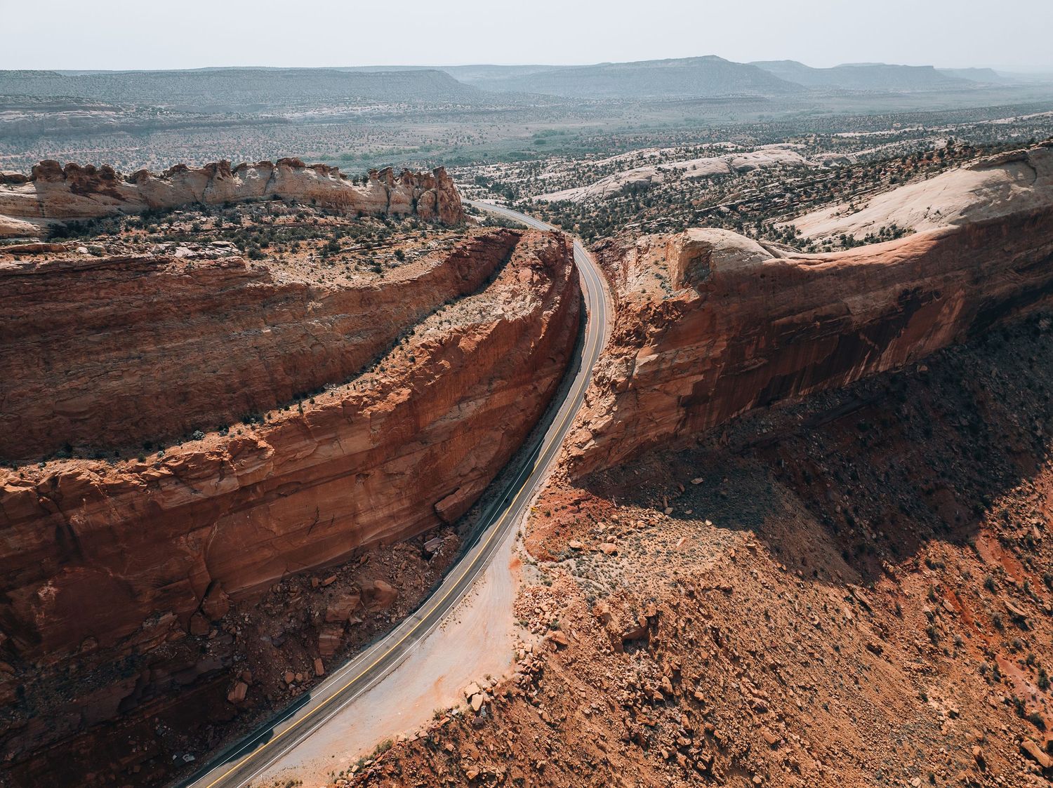 Winding highway road cutting through massive red rock formations in a desert landscape with mountains in the distance.