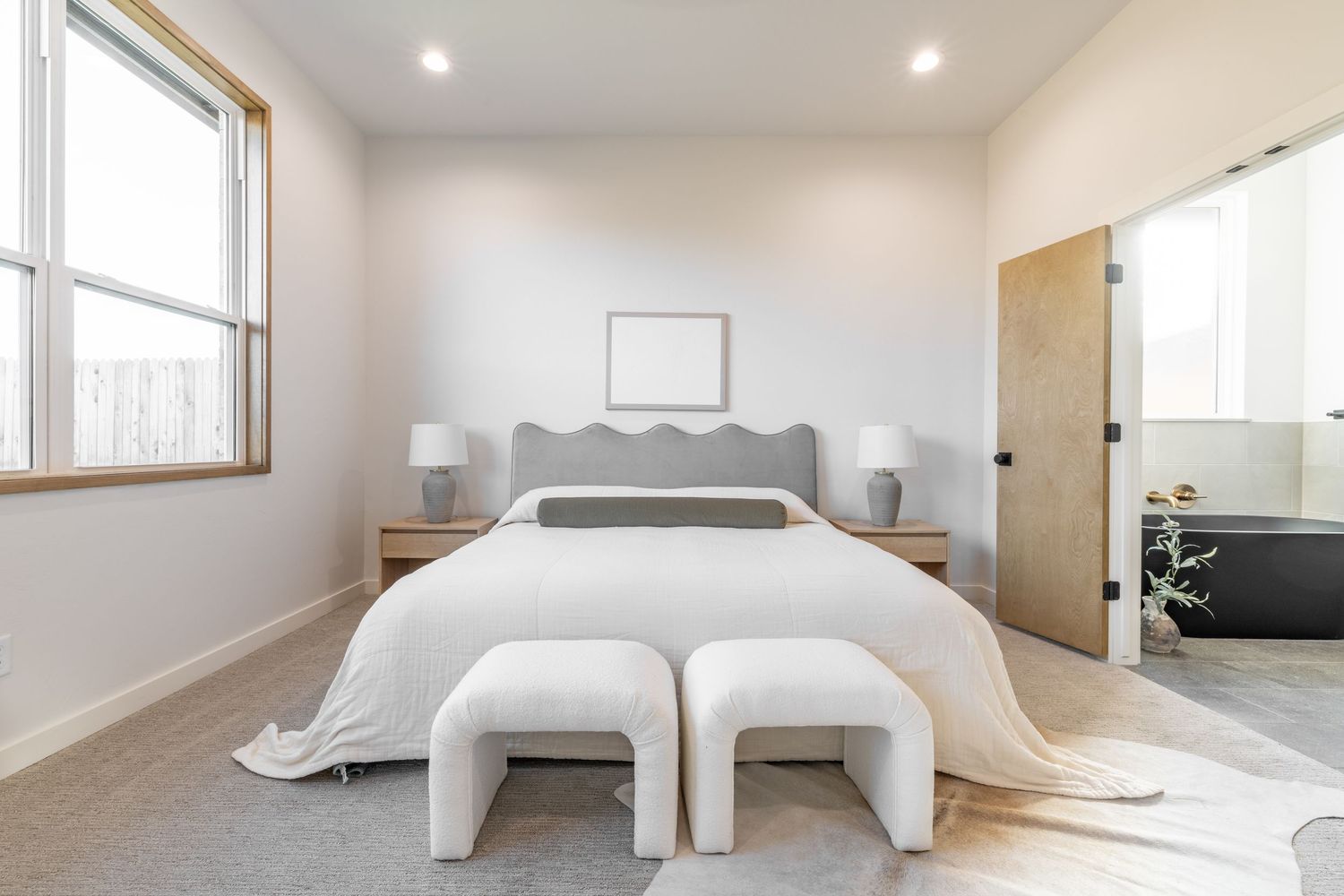 Modern white bedroom interior with gray bed, twin ottomans, and natural light from window in minimalist design style.