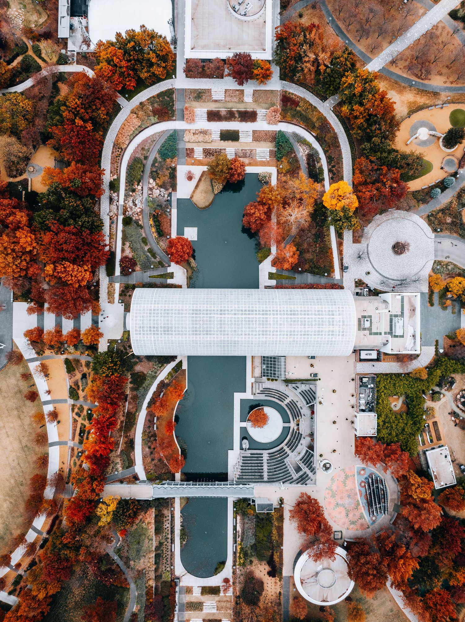 Aerial view of autumn park with geometric architecture, water features and vibrant orange trees surrounding white structures.