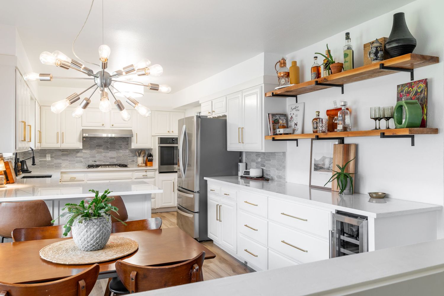 Modern white kitchen with sputnik chandelier, floating wood shelves, marble countertops and stainless steel appliances.