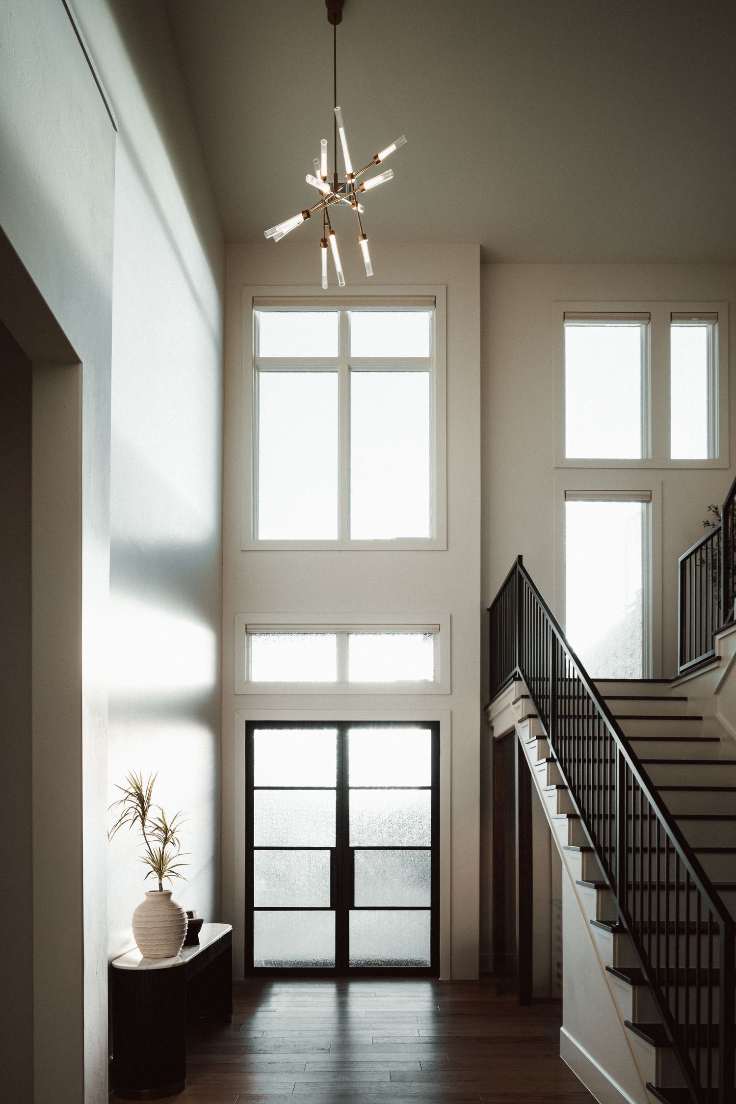 Modern foyer features tall windows, a sleek staircase with black railings, and a contemporary sputnik chandelier in natural light.