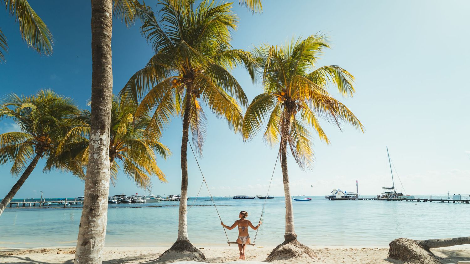 Person enjoying a tropical beach paradise with palm trees and boats anchored in turquoise waters at sunset.