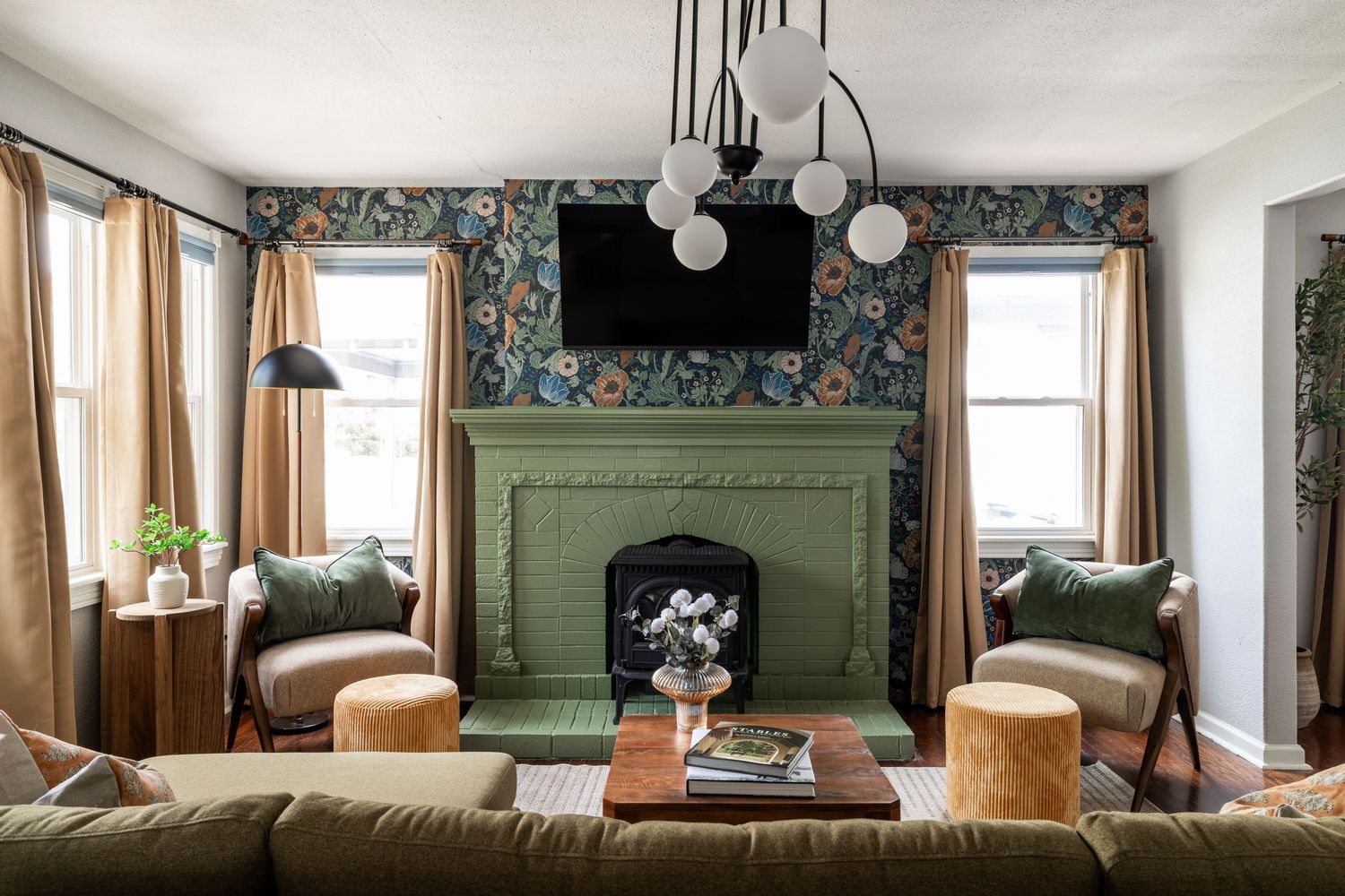 A cozy living room features sage green built-in cabinets, floral wallpaper, and warm natural light from multiple windows.