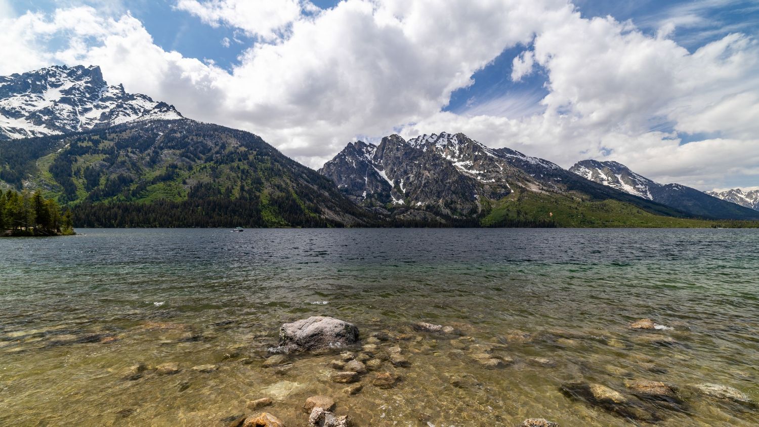 Clear mountain lake with rocky shore reflects snow-capped Grand Teton peaks under puffy white clouds in Wyoming.